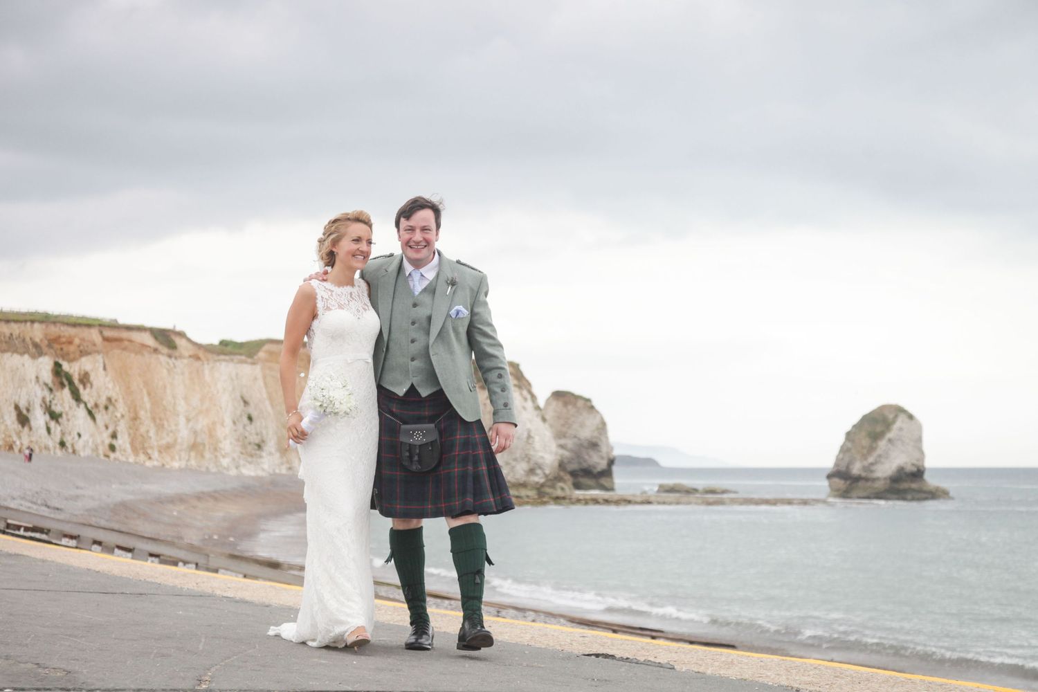 A couple walks along a scenic coastal beach with white cliffs and ocean views in the background.