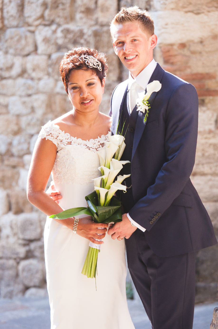Joyful wedding couple poses with white calla lilies against stone wall.