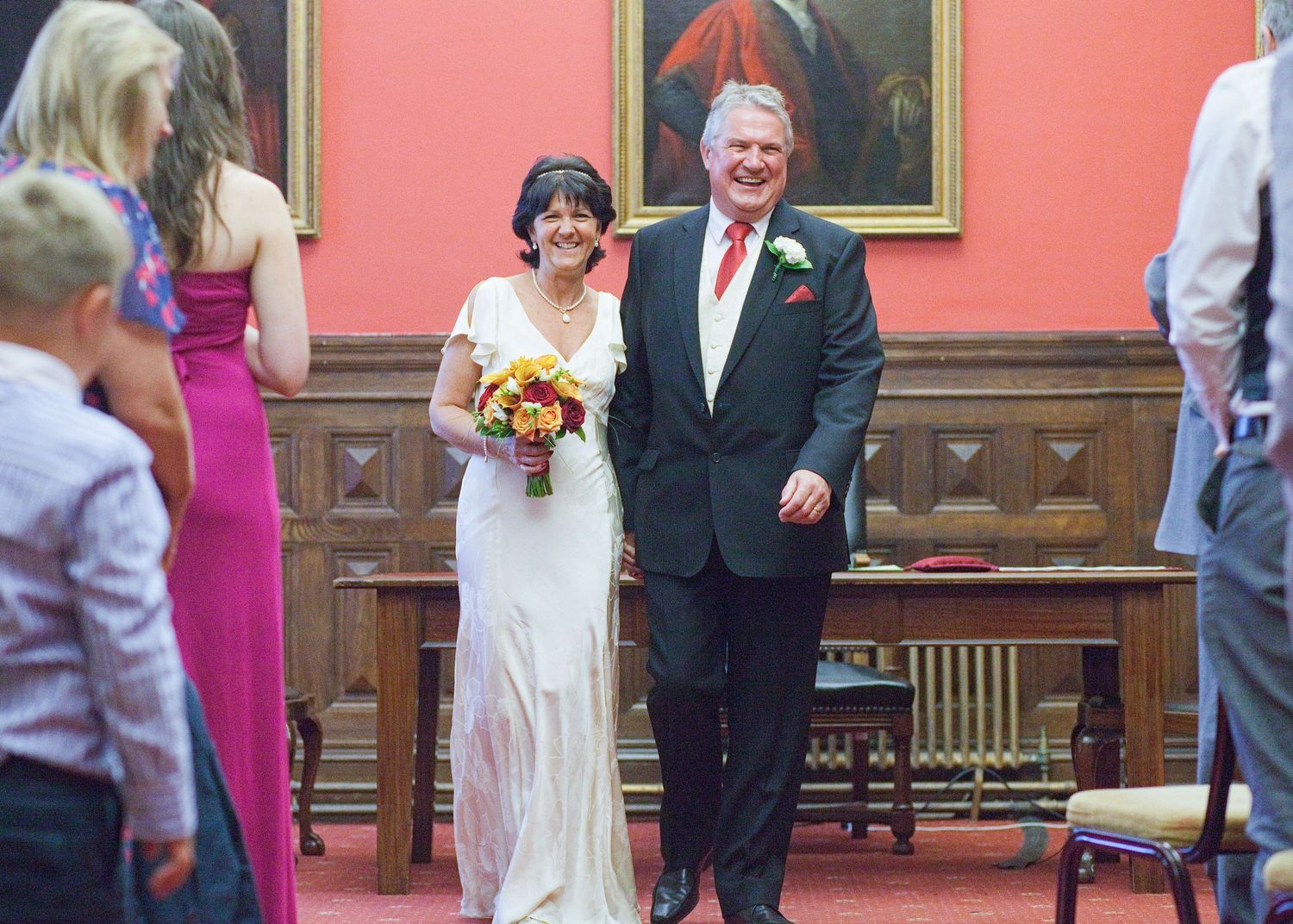 A wedding couple walking down an aisle in a room with red walls and ornate framed paintings.