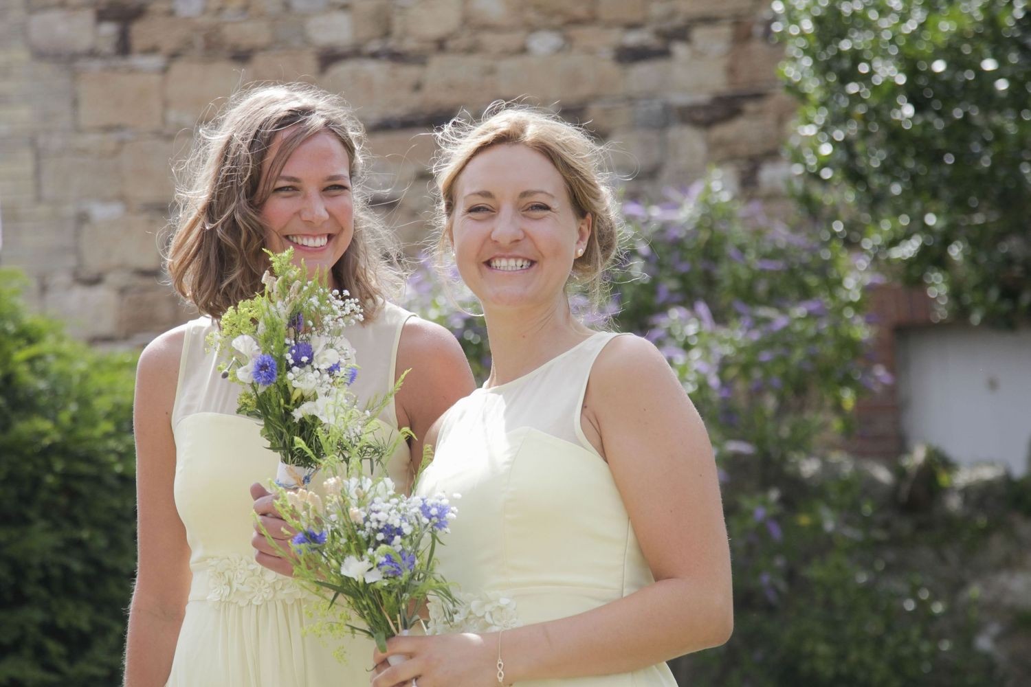 Wedding party members share joyful moments near a rustic stone wall.