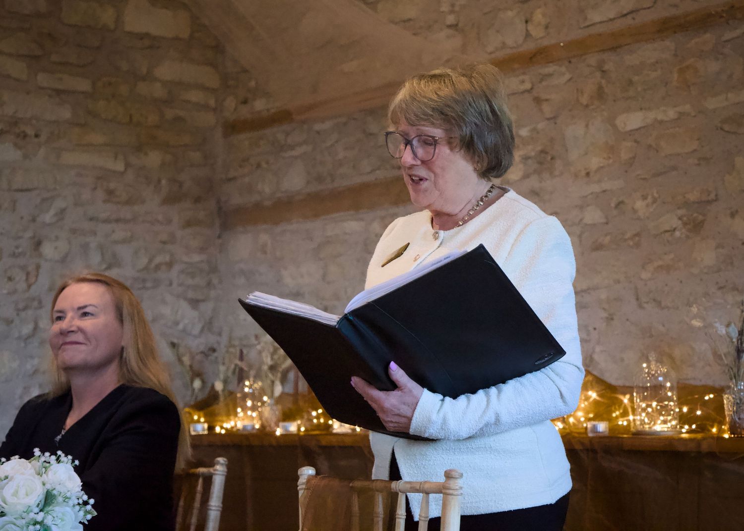 A woman in white reads from a large black book during a ceremony in a stone room with glowing candles.