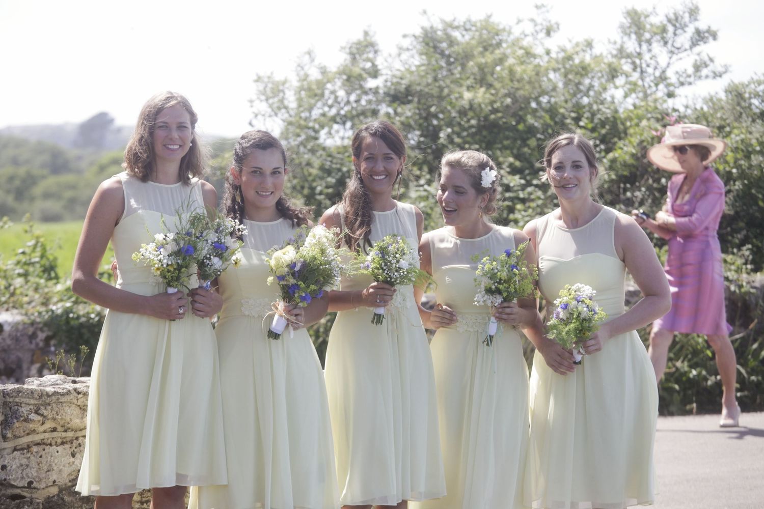 Bridesmaids in cream-colored dresses holding bouquets pose together outdoors.