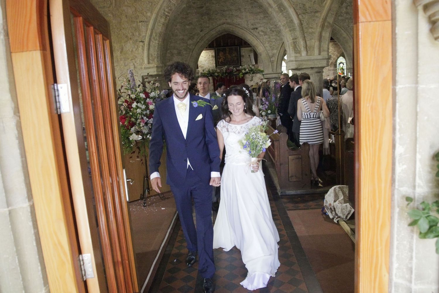 Wedding procession walking through arched church corridor decorated with flowers.