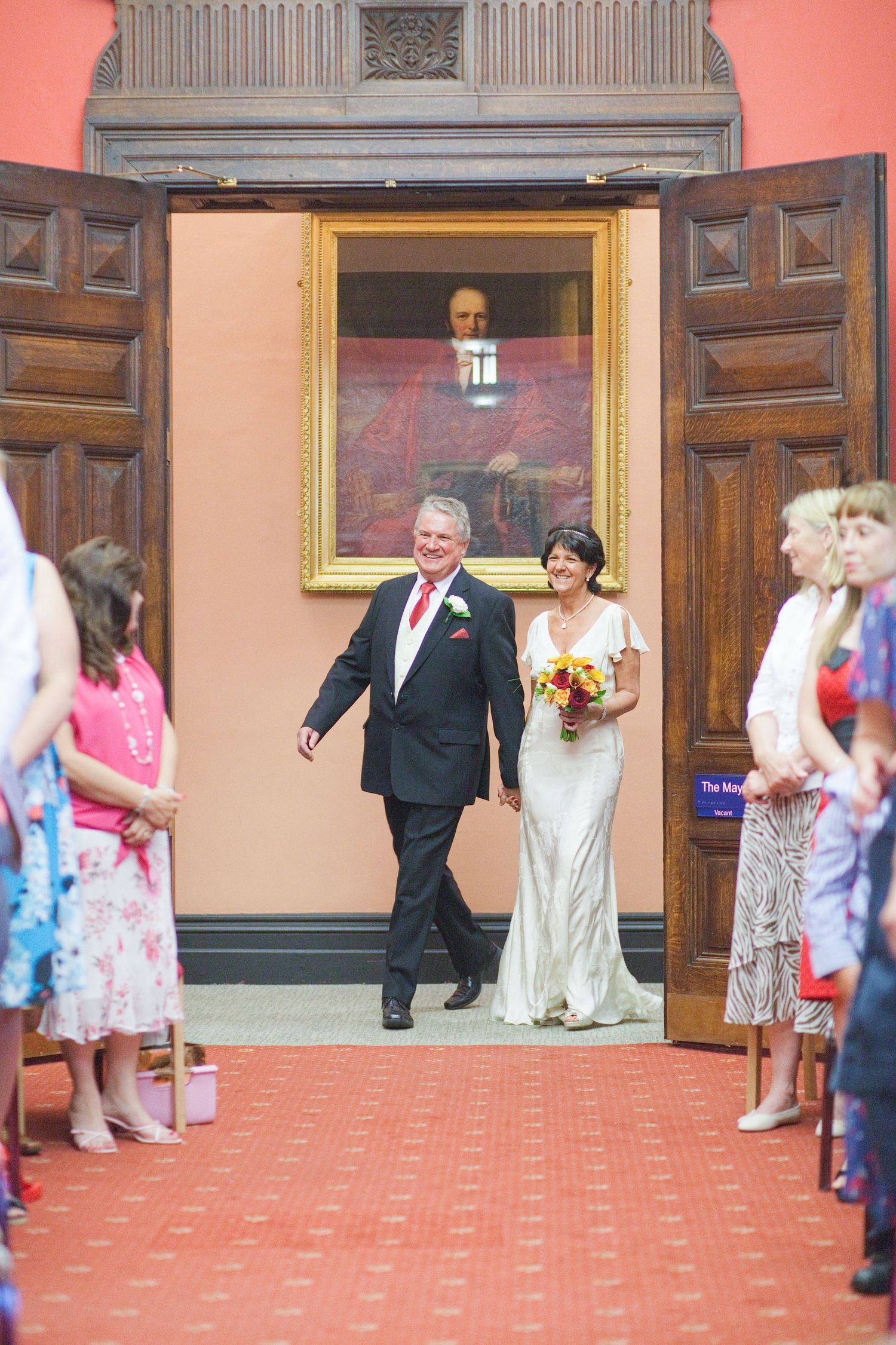 A wedding procession walks down a carpeted hallway with wooden doors and coral walls.