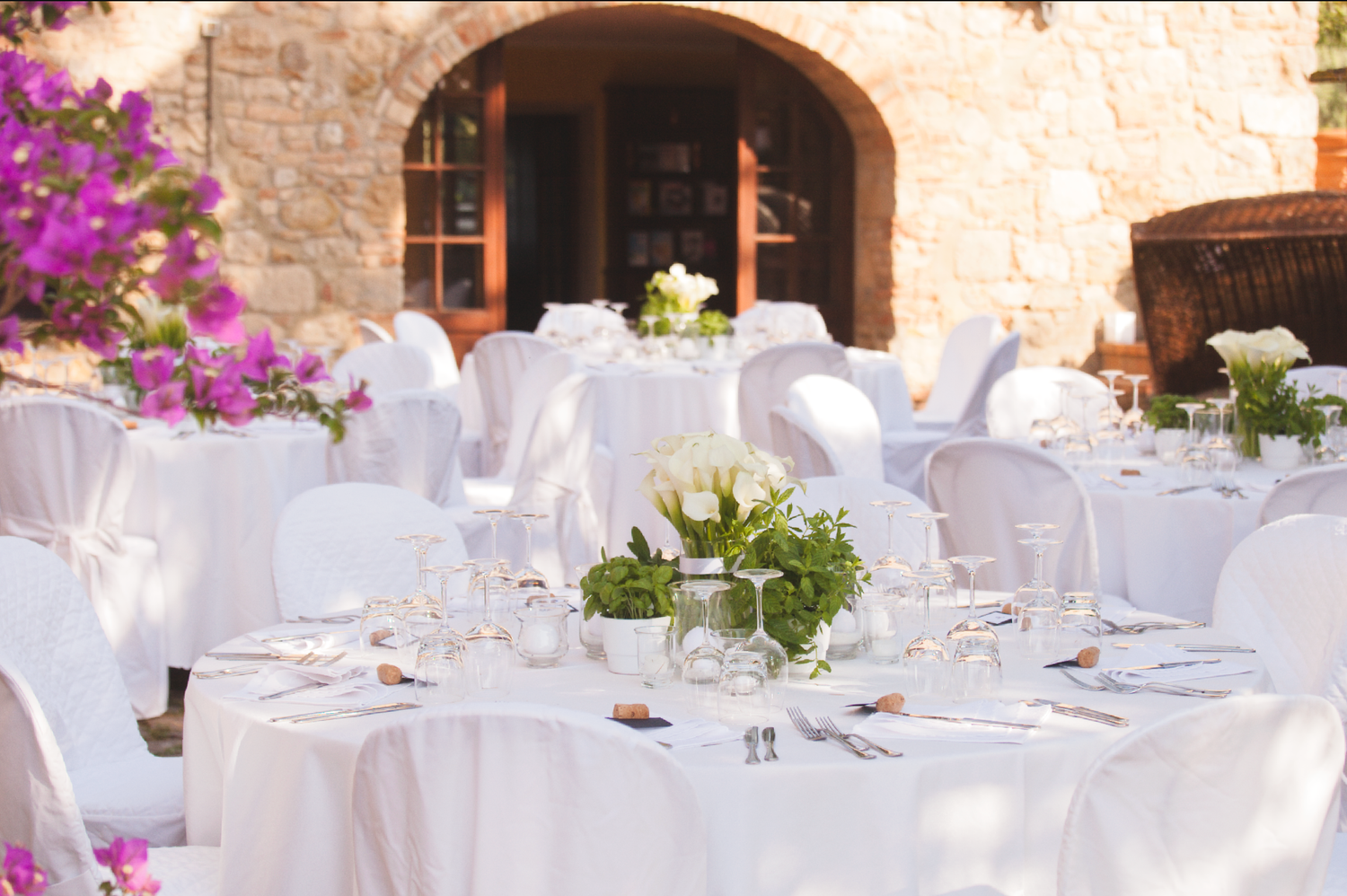 Elegant outdoor wedding reception setup with white tablecloths and floral centerpieces under a stone archway.