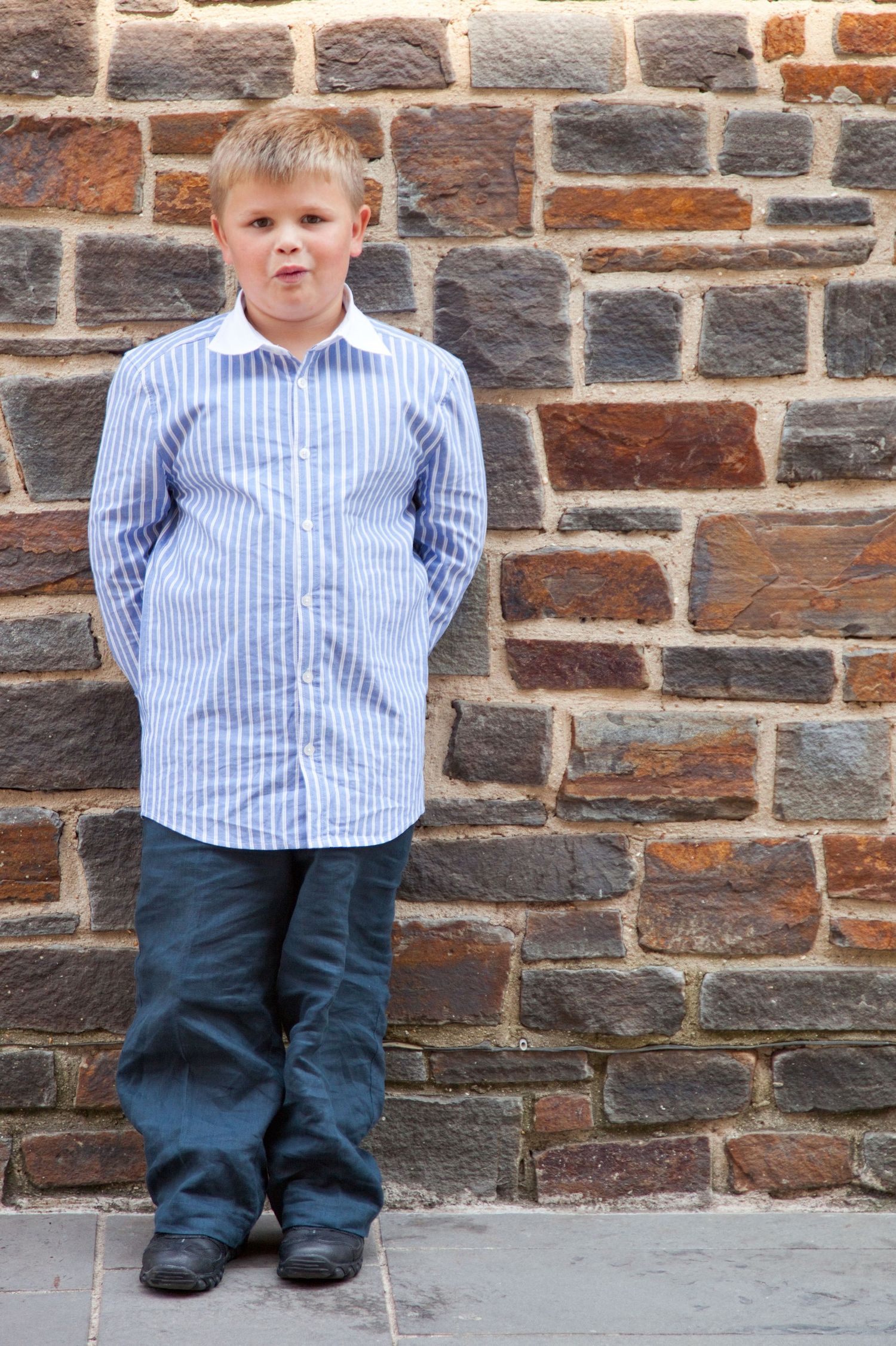 A small child wearing a blue striped shirt and dark pants stands against a rustic brick wall.