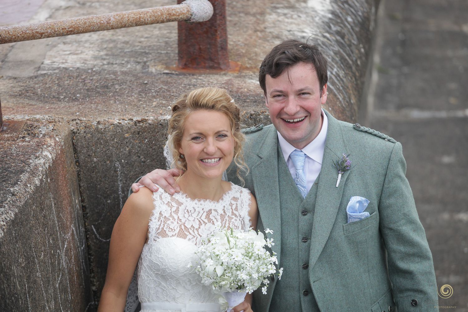 Wedding couple poses along a rustic stone wall by the seaside.