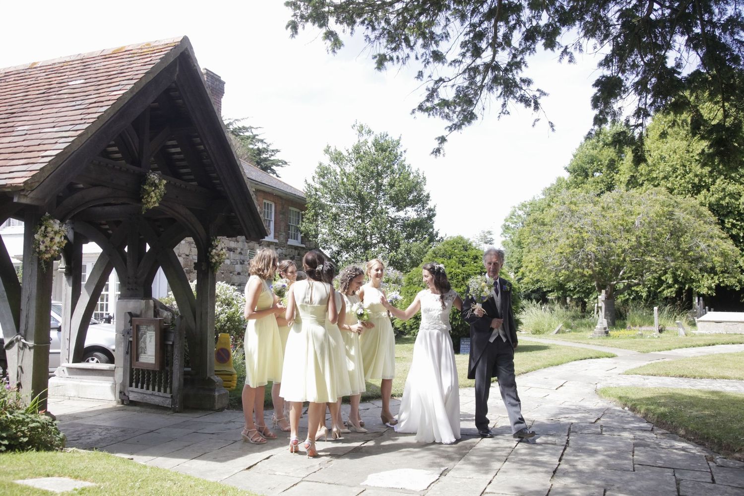 Wedding party gathered under a wooden lych gate outside a traditional church.