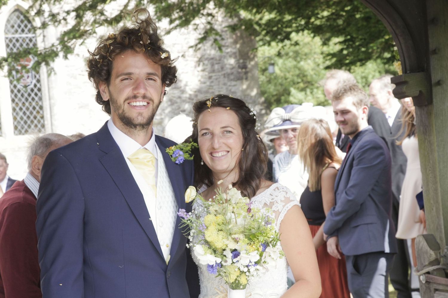 Wedding party gathers for photos outside stone church on sunny afternoon.