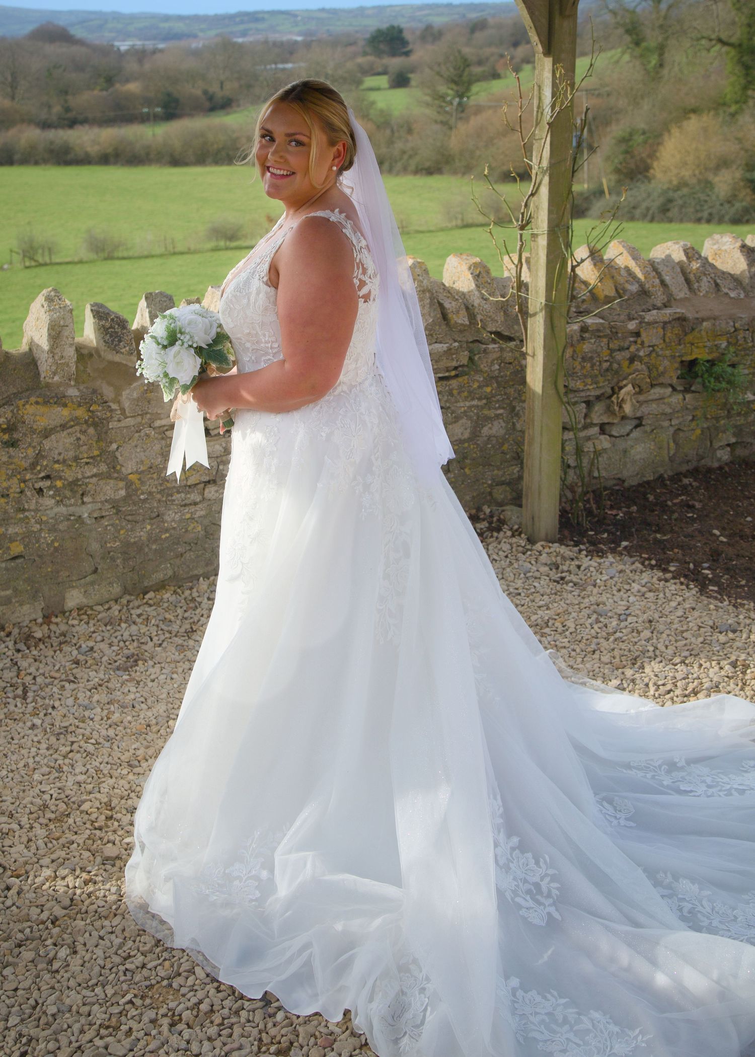 Elegant white wedding dress with lace details and flowing train photographed against stone wall backdrop.