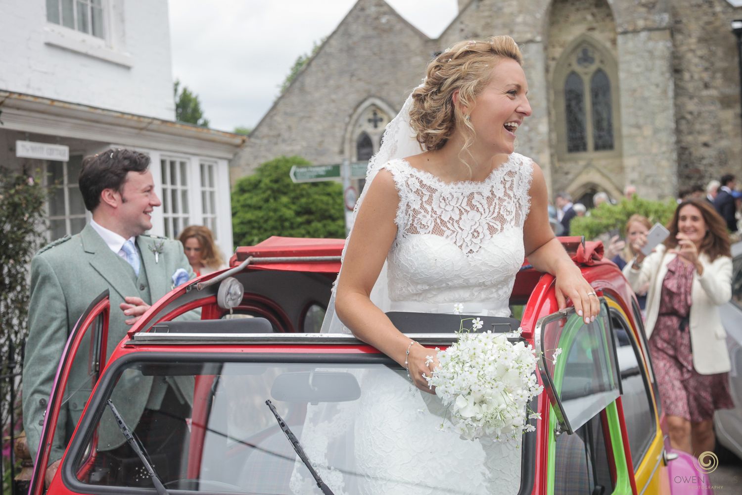 Bride waves her boquet from the sunroof of a colourful Citroen 2CV during a wedding photography phoot shoot after the church service.