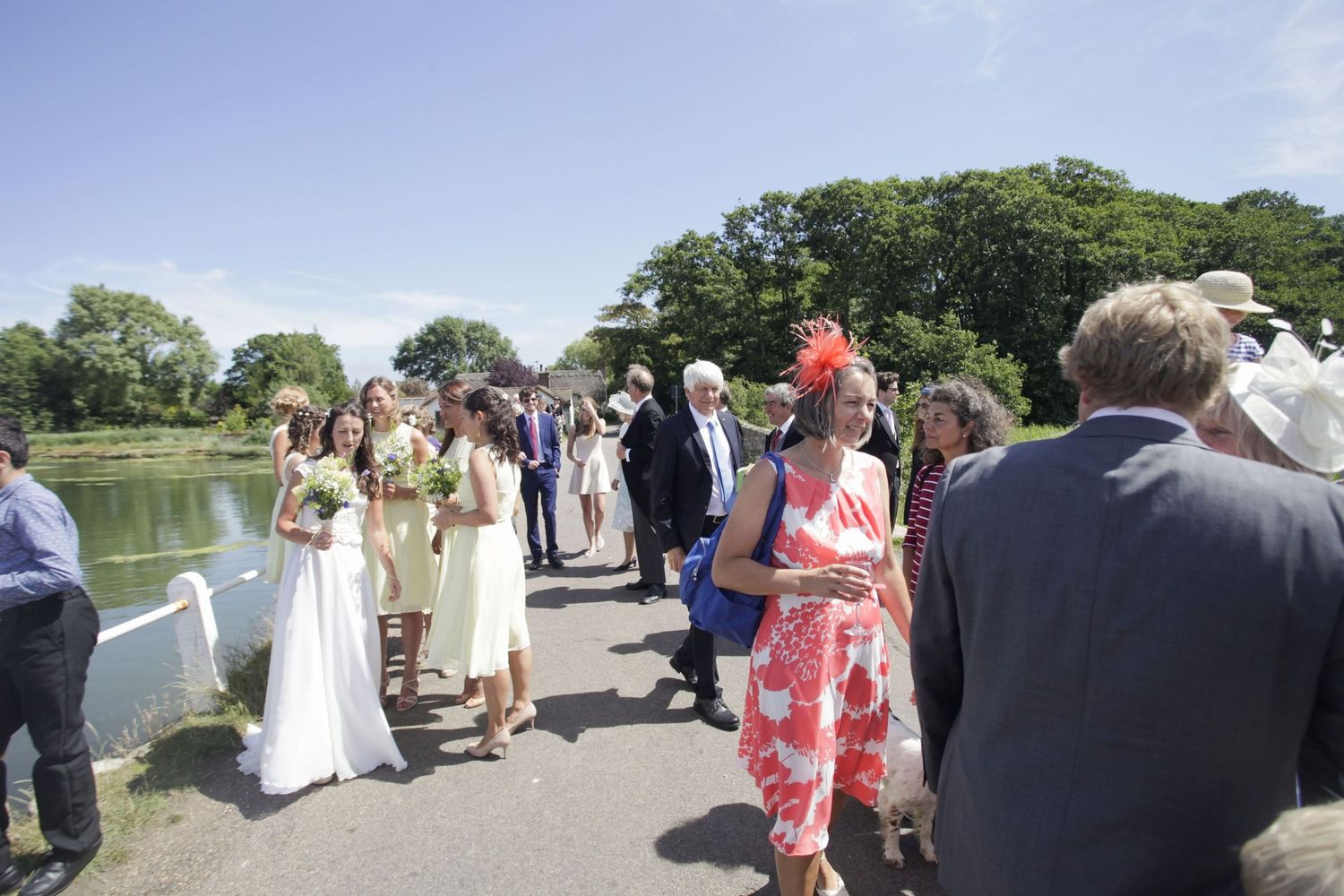 Celebration guests in formal attire gather along a sunny lakeside path.