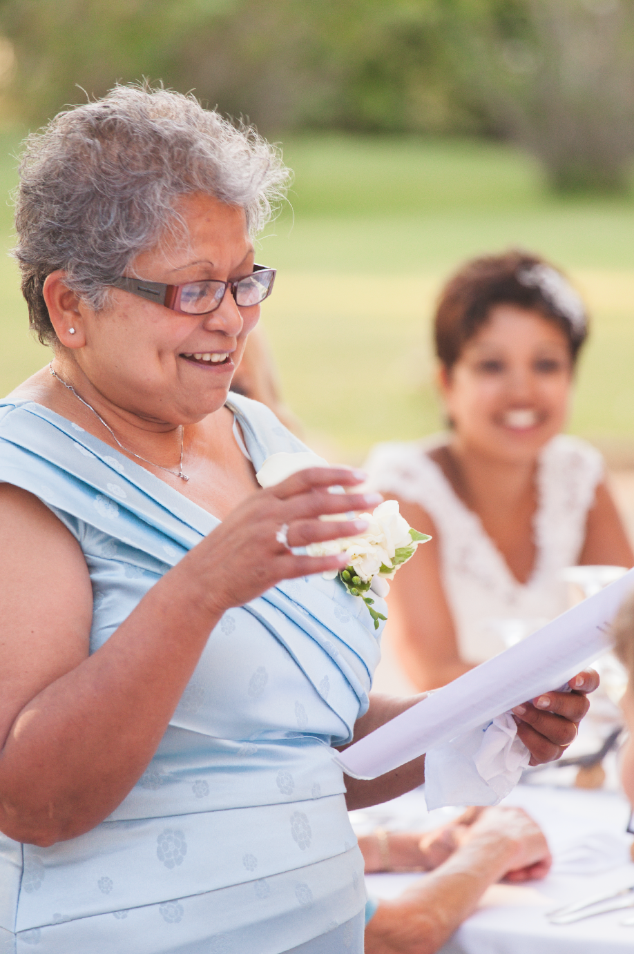 A guest in a light blue dress reads from papers during an outdoor wedding celebration.