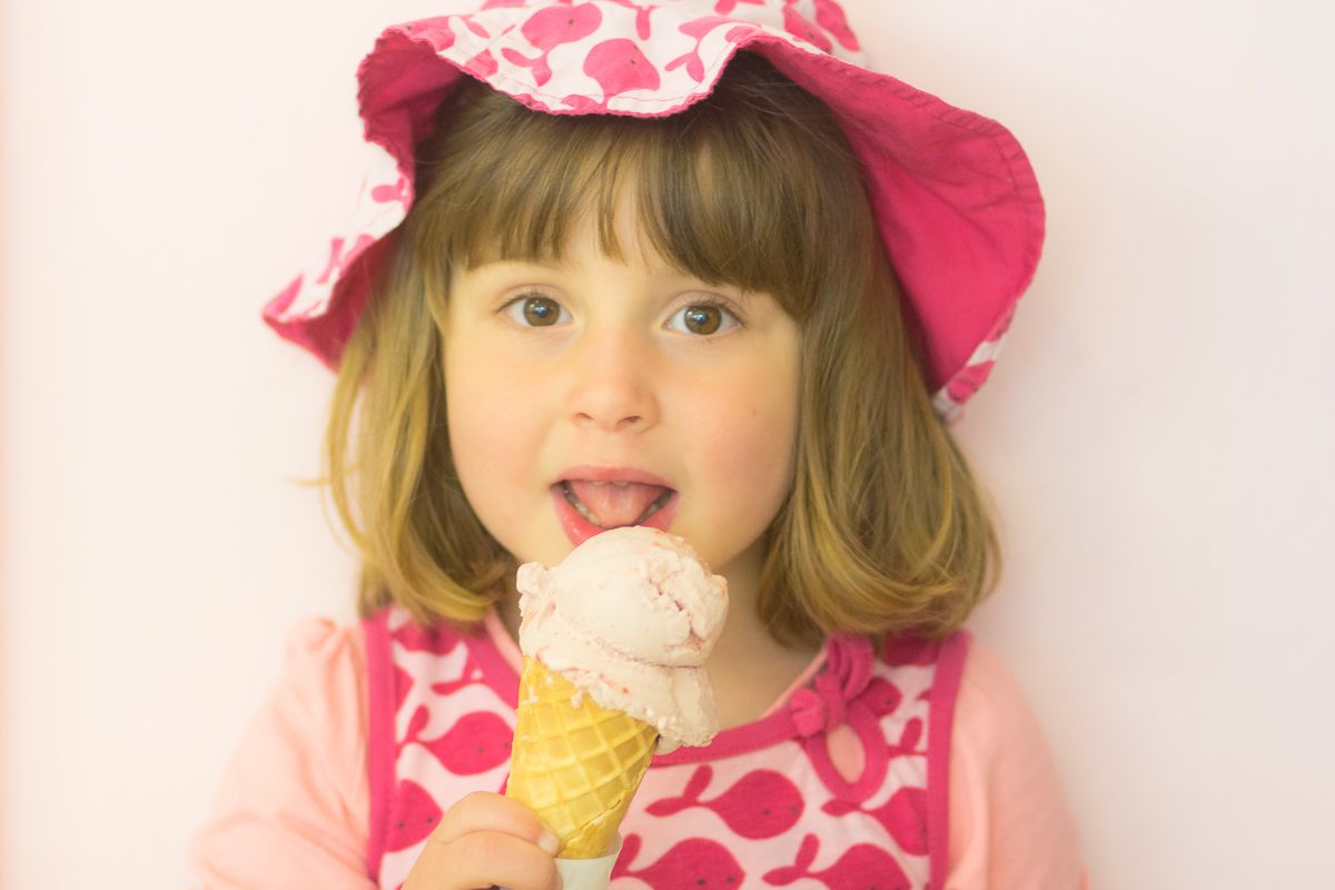 A child in a pink patterned dress and matching sun hat enjoys licking an ice cream cone against a white background.