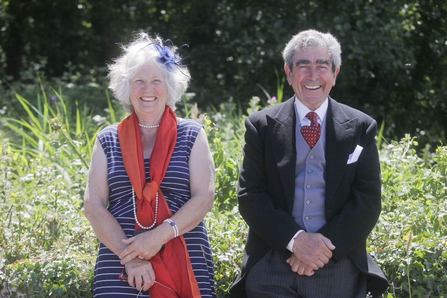 Couple sitting together outdoors with red and navy accessories against green foliage.