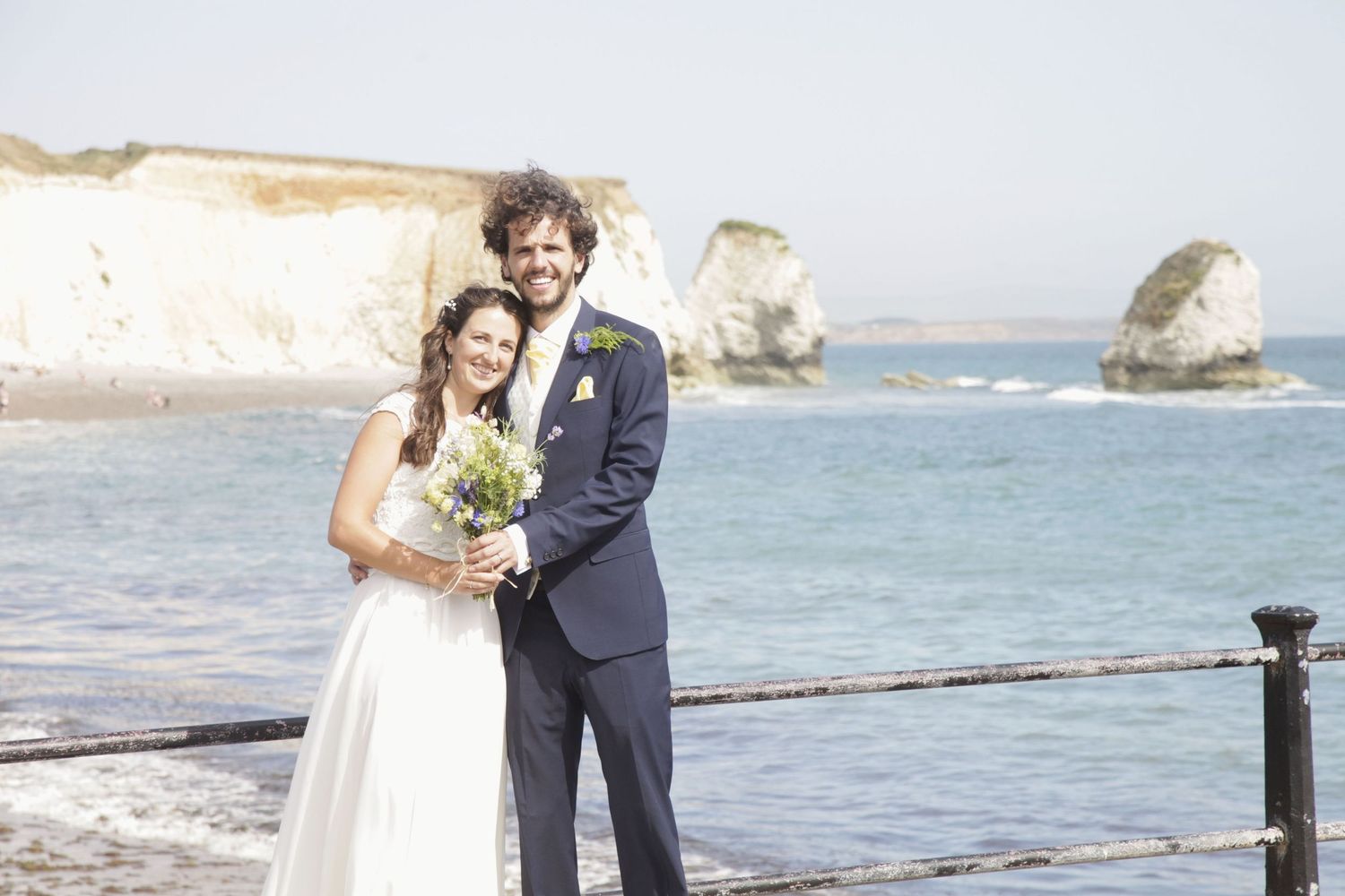 Wedding couple posing on scenic coastal path with ocean and rock formations in background.