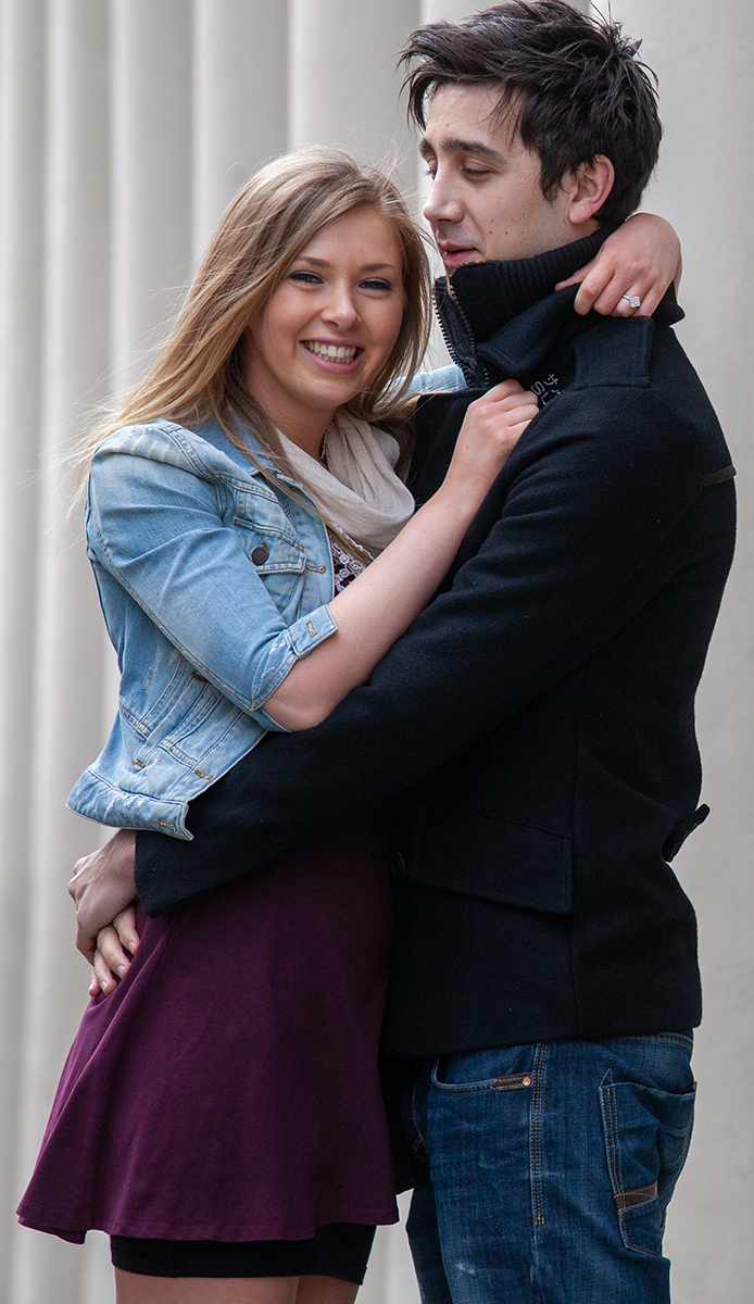 A couple embraces casually while posing against a white column backdrop.