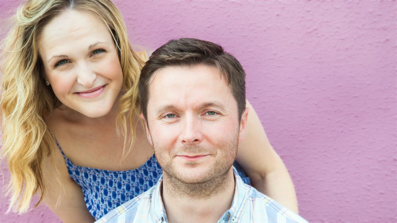 A couple smiles warmly while posing together against a bright pink wall.