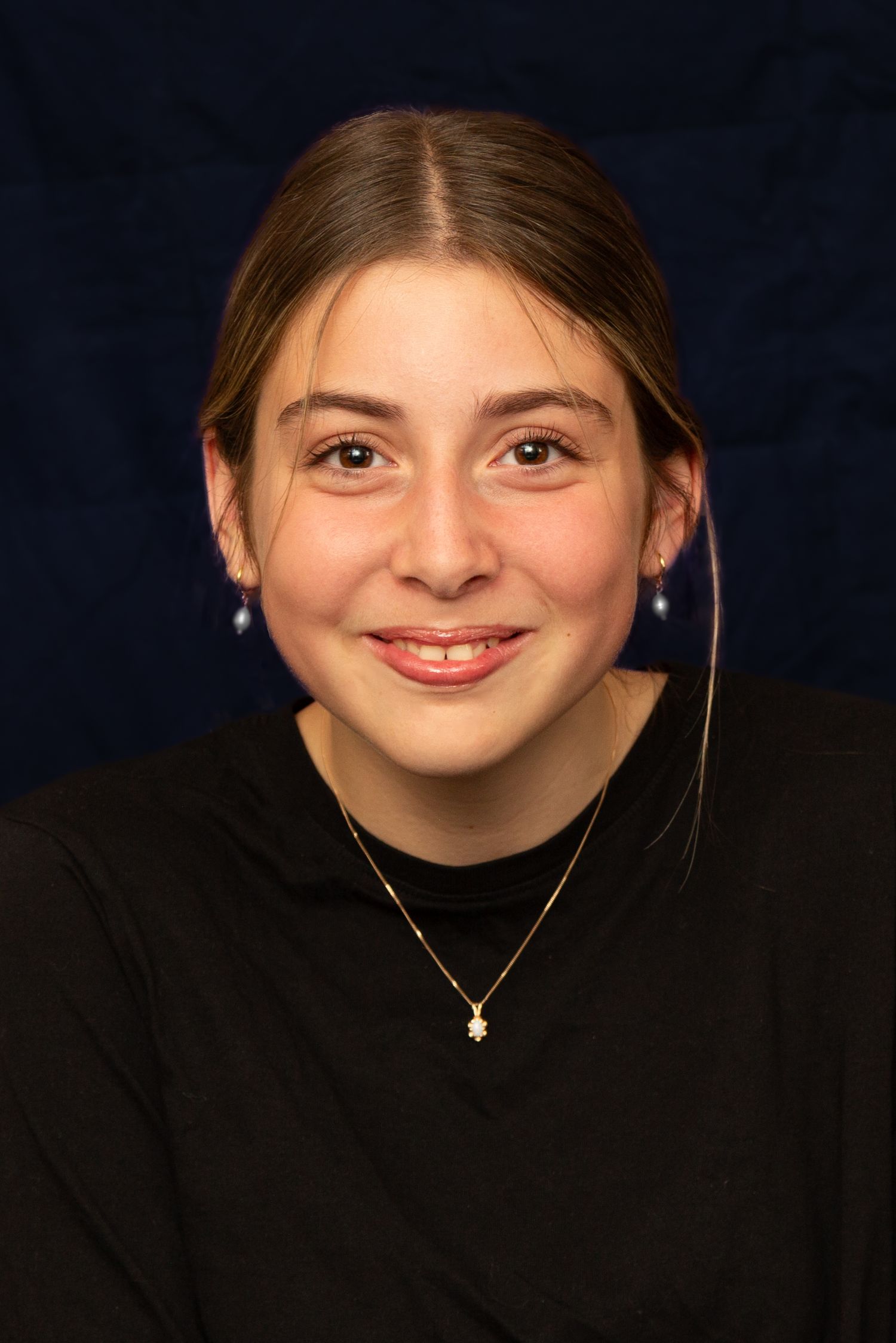 Portrait of someone in a black sweater with pulled back hair, delicate necklace and earrings against dark background.