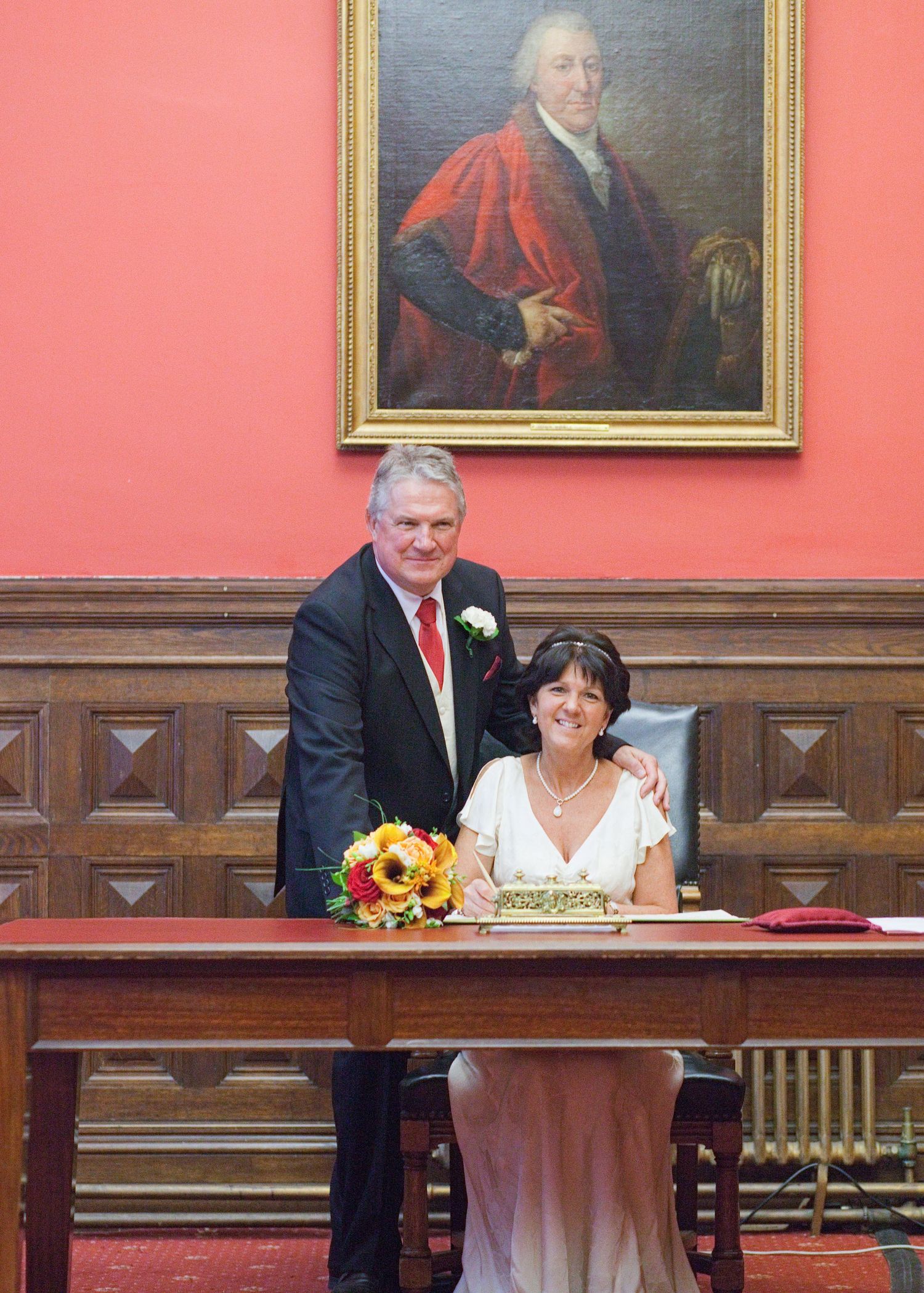 Wedding ceremony in historic room with portraits on coral walls and wooden paneling.