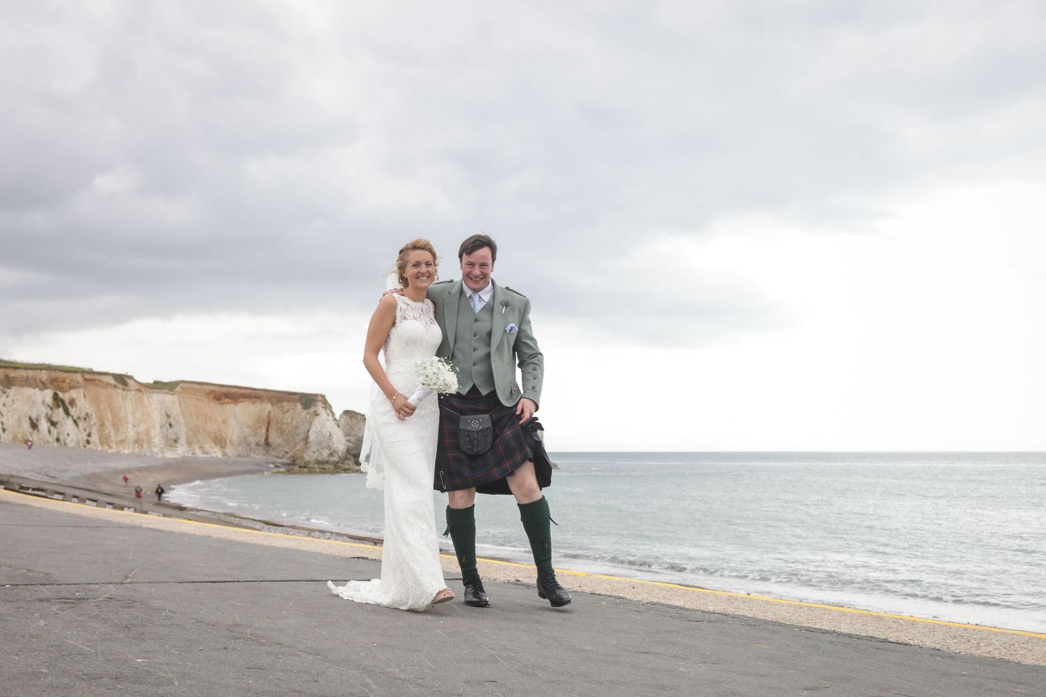 Wedding couple strolls on a rocky beach with dramatic coastal cliffs in the distance.