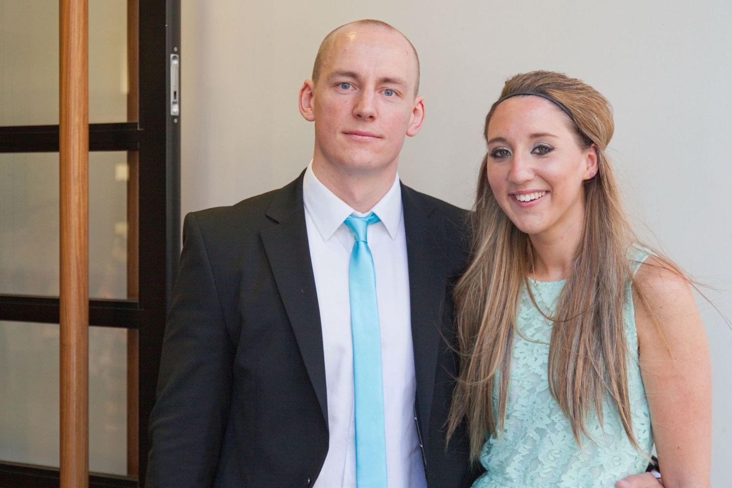 Formal portrait of couple in mint blue and black attire against white wall.