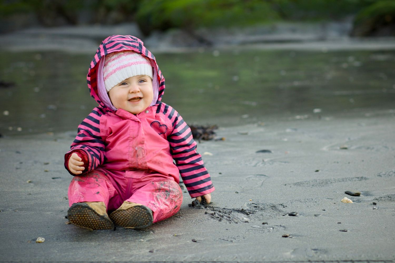 A small child in a pink snowsuit sits and plays on wet pavement during winter weather.