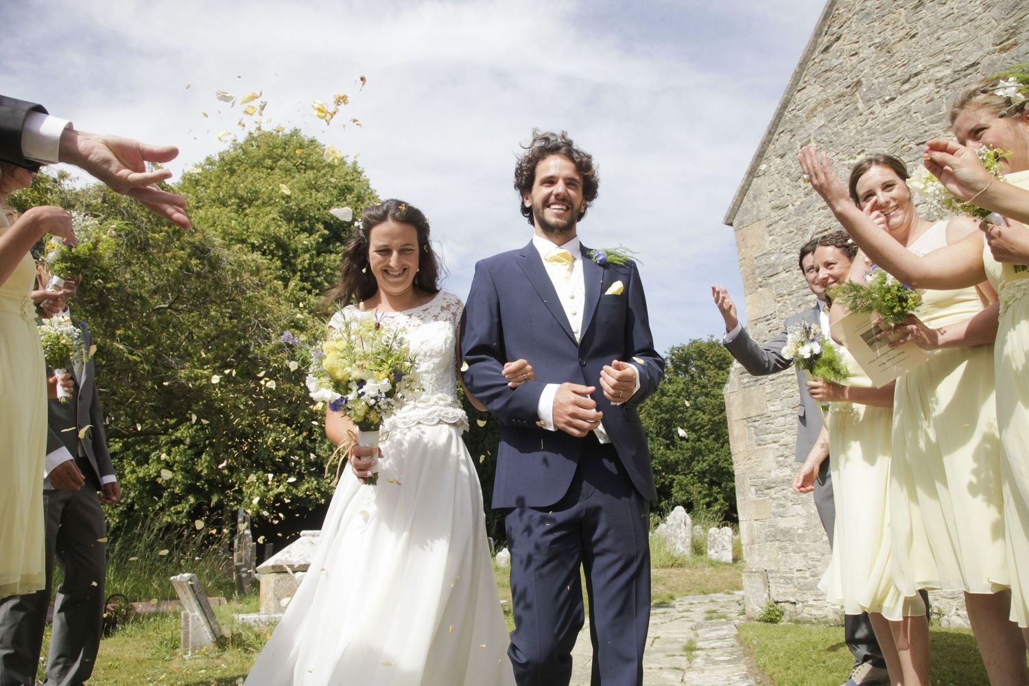 Wedding guests throw confetti and celebrate outside a stone church on a sunny day.