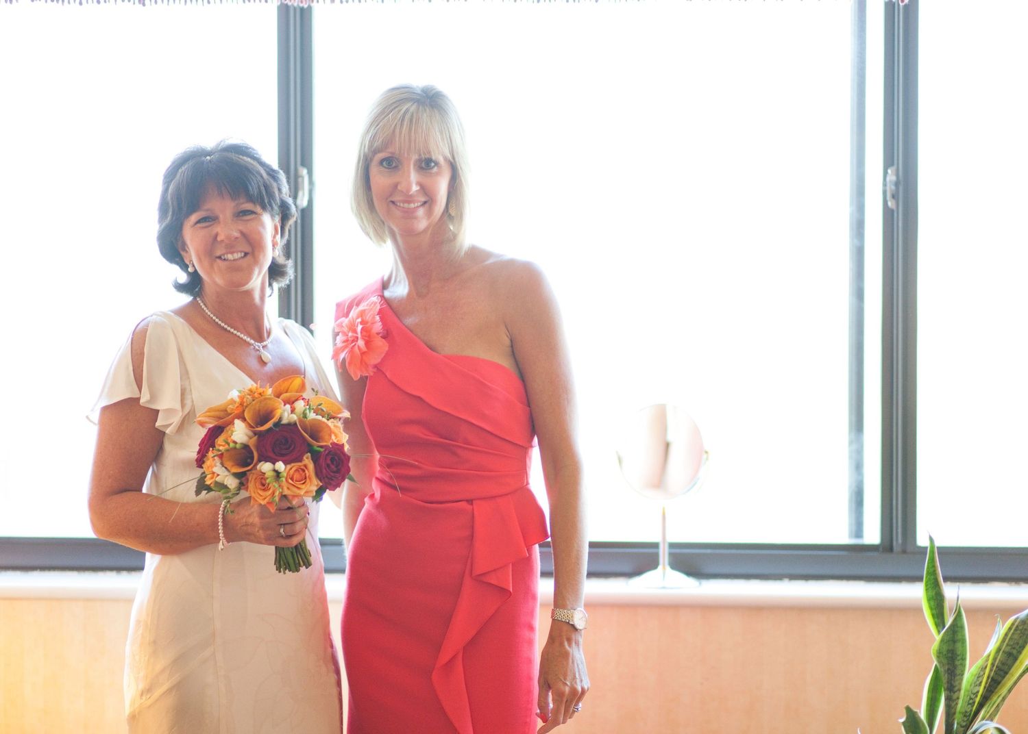 Two people in formal dresses pose with a colorful bouquet near large windows.
