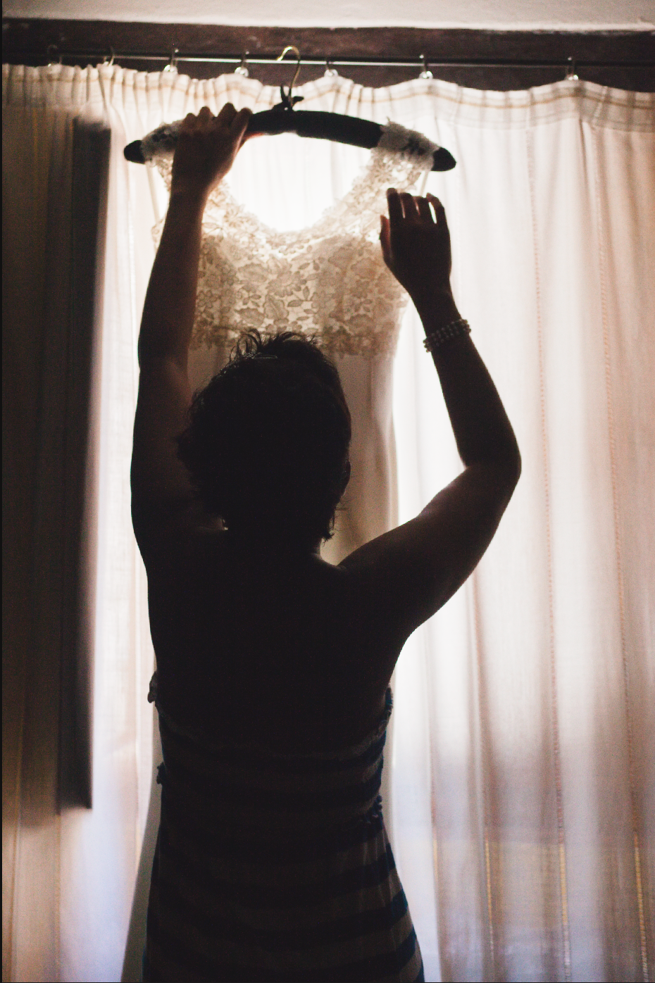 Silhouette of wedding dress hanging against white curtained window.