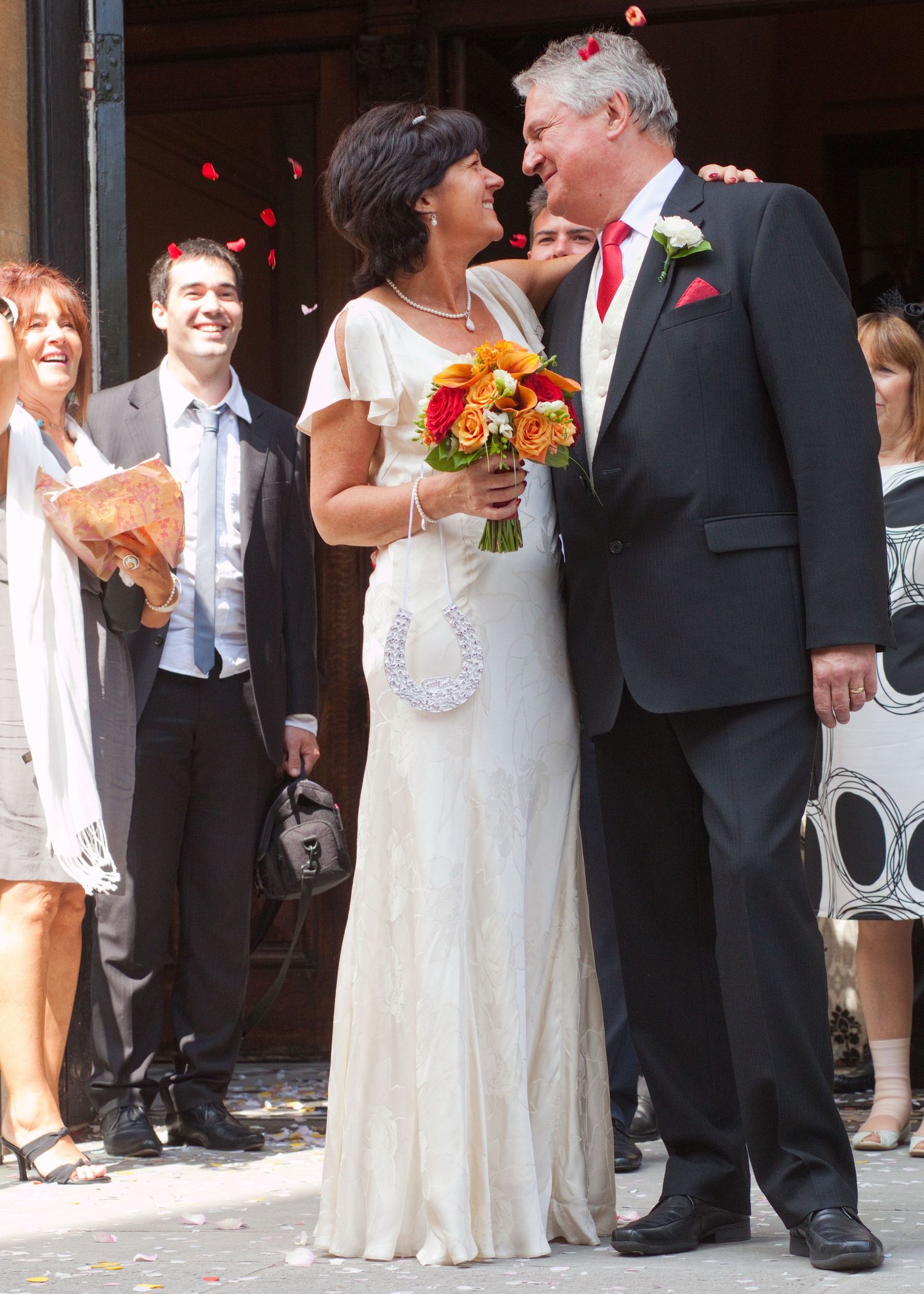 A newlywed couple embracing outside while rose petals fall around them.