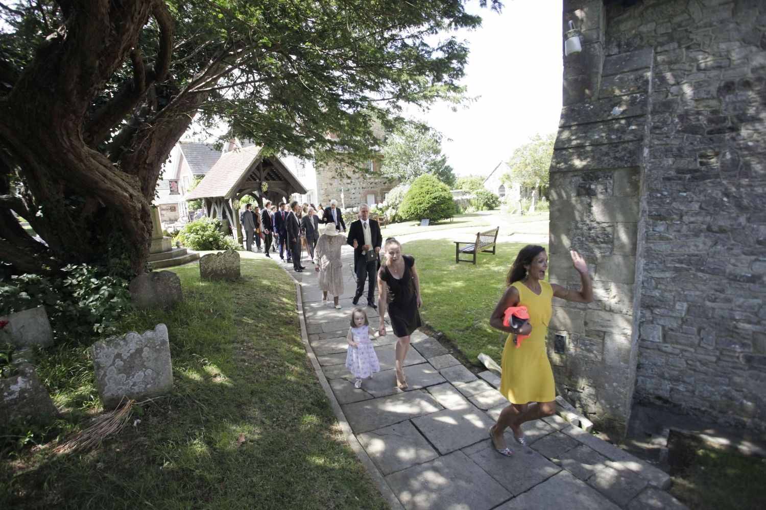 Wedding guests walking along a garden path outside a stone church on a sunny day.