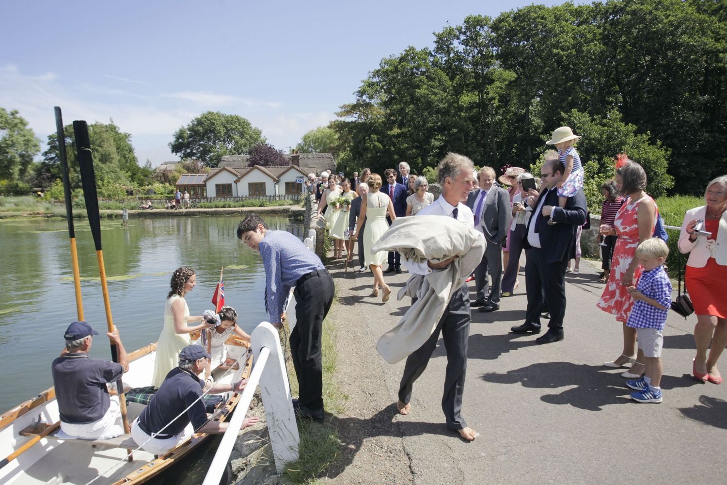 Guests in summer attire gather along a lakeside path watching a boat ceremony.