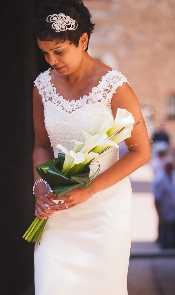 A bride in a white lace dress holds a bouquet of calla lilies against a stone backdrop.