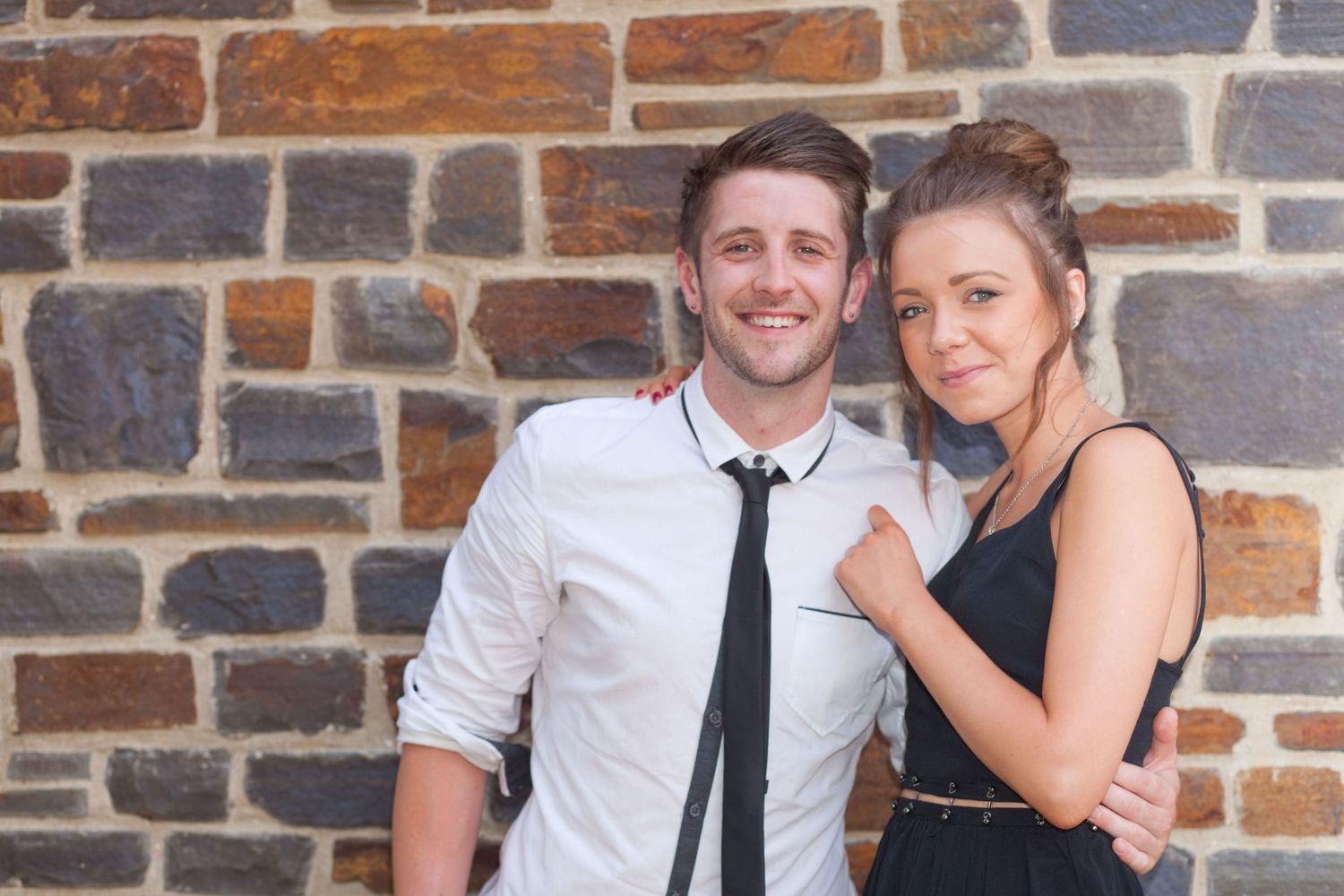 A couple leans against a stone wall while smiling during a casual photo shoot.