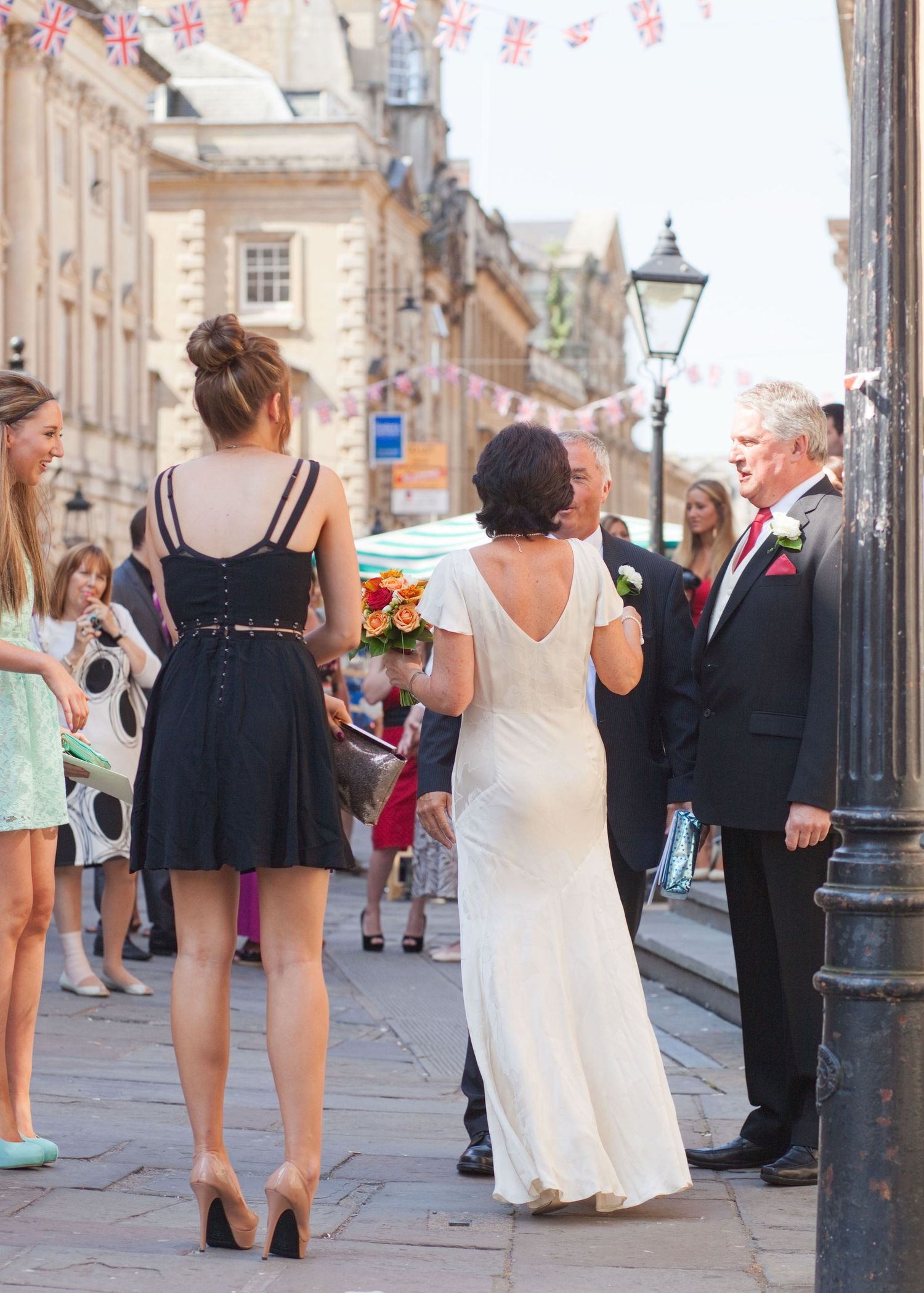 Wedding guests and couple walking along historic street with bunting decorations and classic architecture.