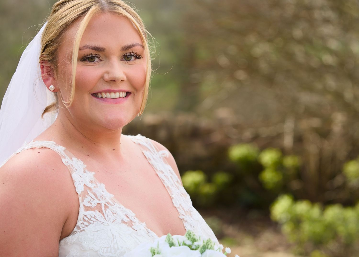 A bride in a white lace wedding dress poses outdoors with a bouquet during a sunny day.
