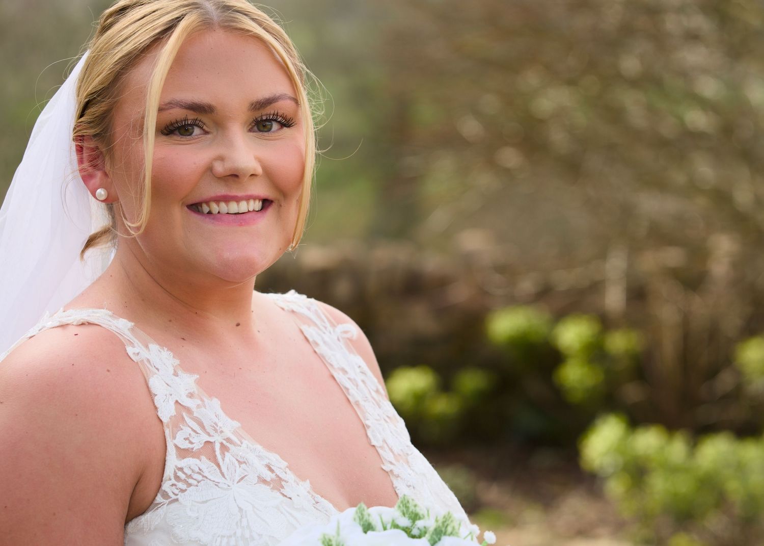 A bride wearing a lace wedding dress smiles radiantly in an outdoor garden setting.