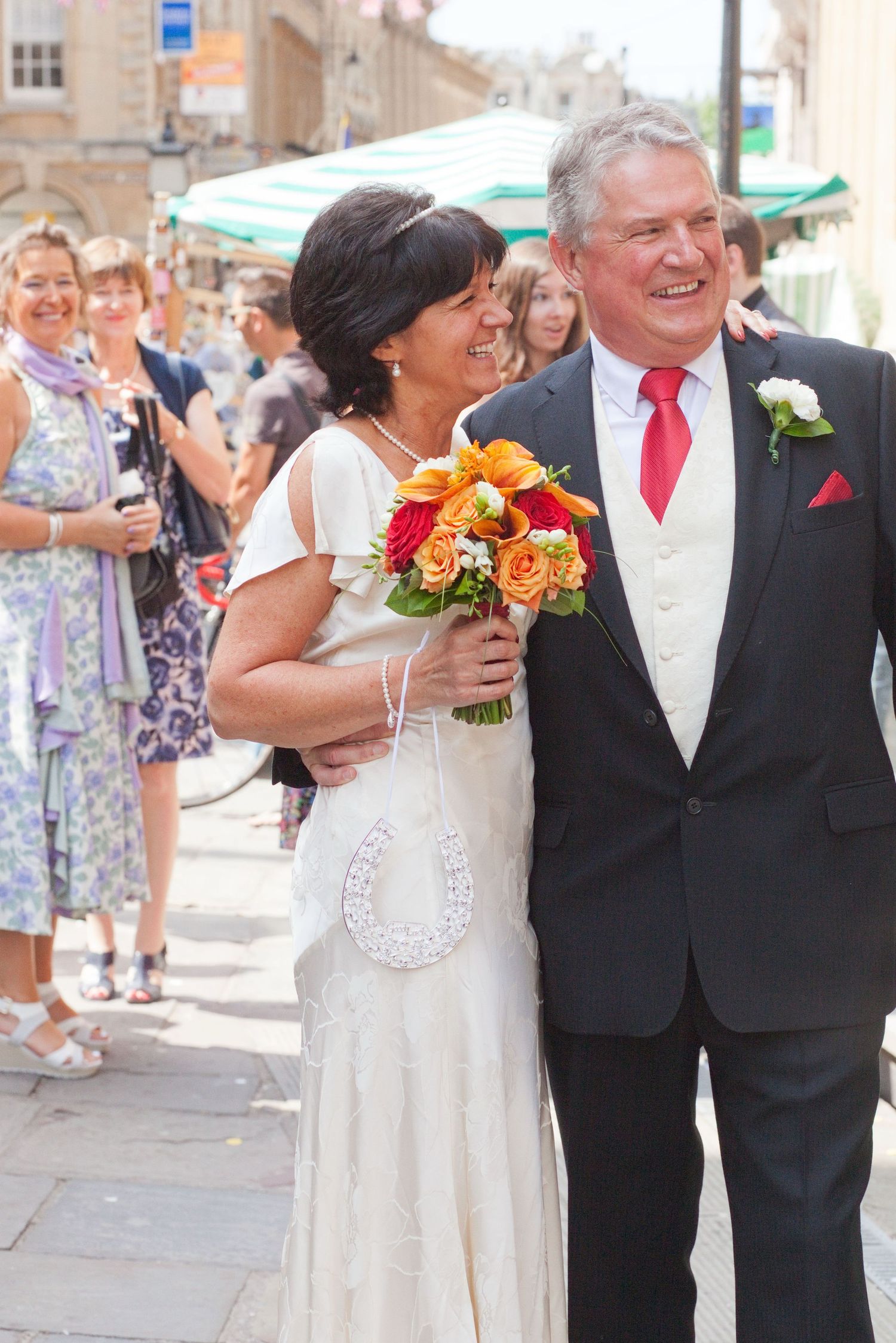 Wedding party poses together on a sunny street with guests in white and formal attire.