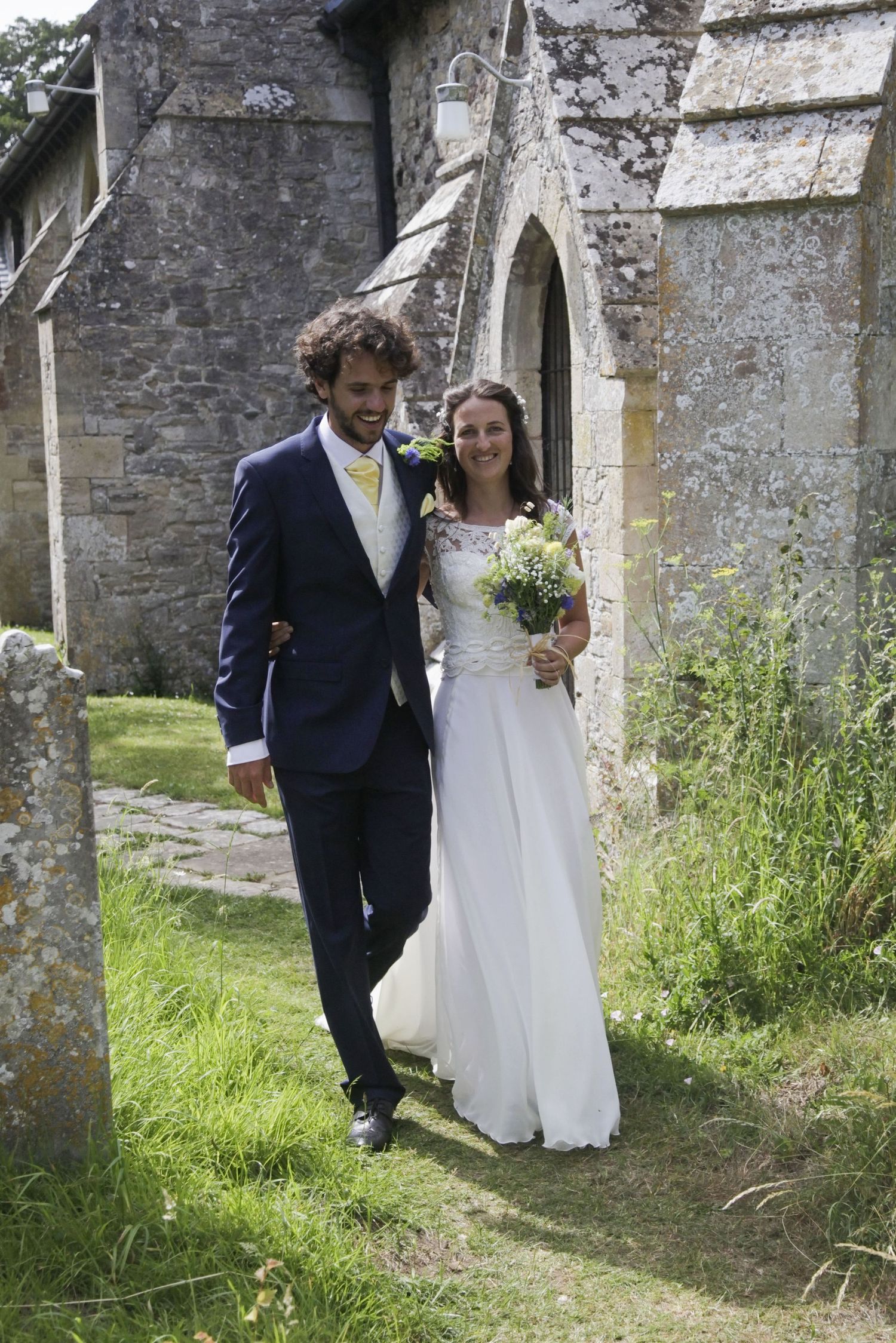 Newlyweds walk together along a stone wall path at a historic church.