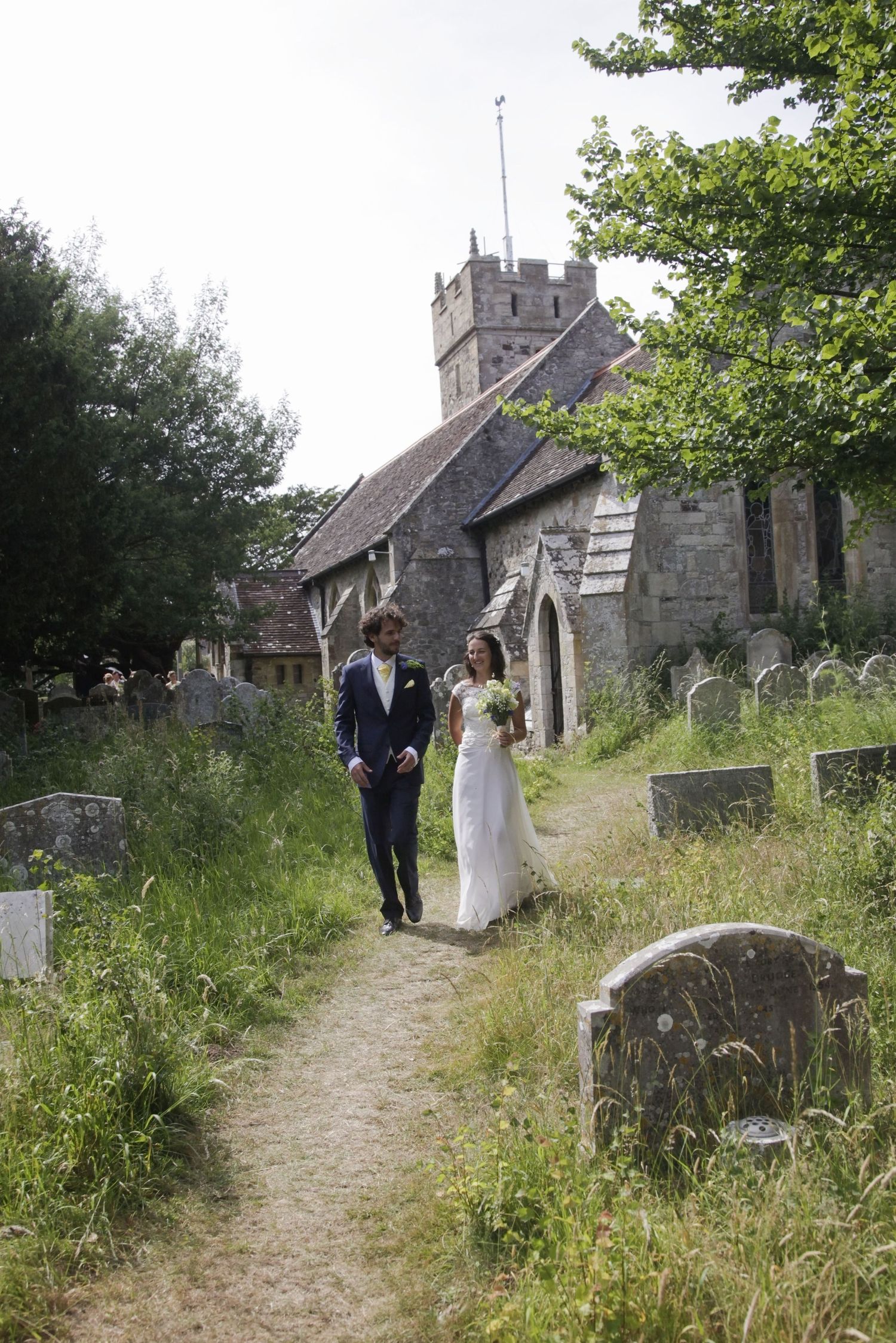 A wedding couple walks through an old church graveyard with headstones and overgrown grass.