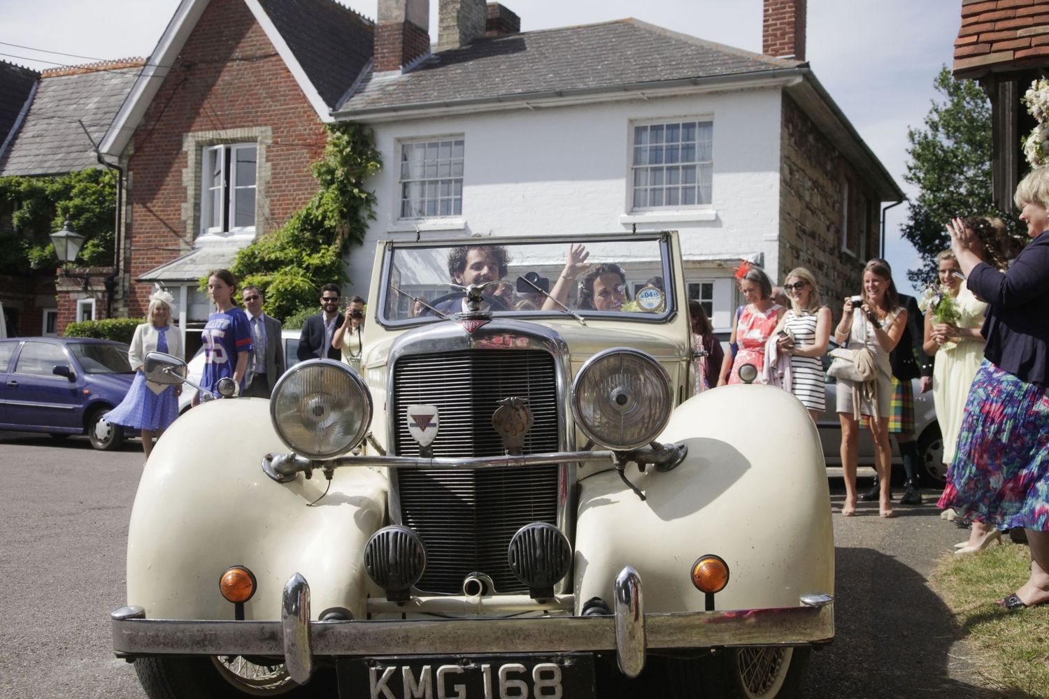 Wedding party gathered around classic cream-colored vintage automobile near country house.