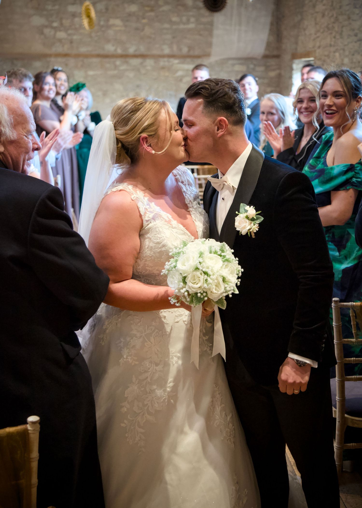 Wedding couple shares a kiss while guests applaud in a beautifully lit stone venue.