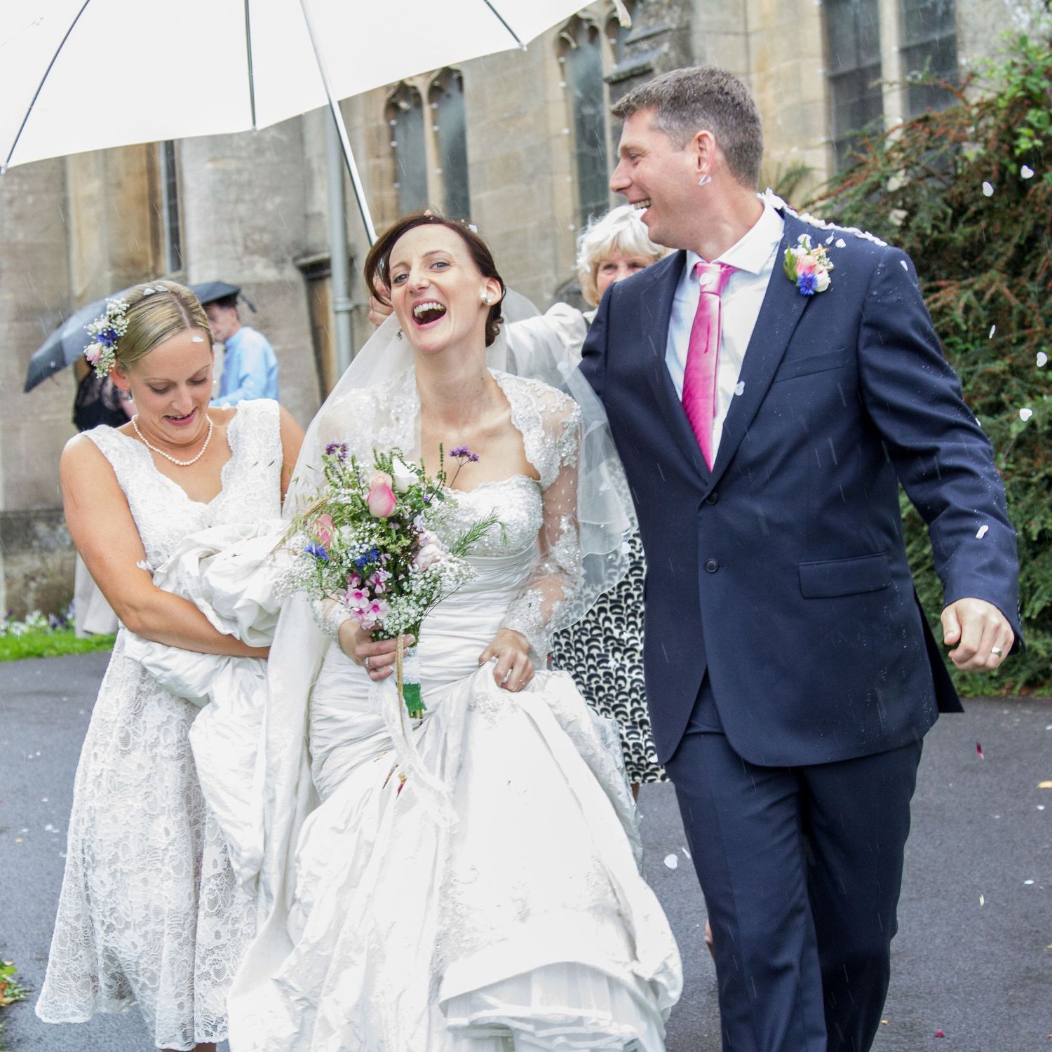 Wedding party laughs together while holding white umbrellas on a cloudy day.