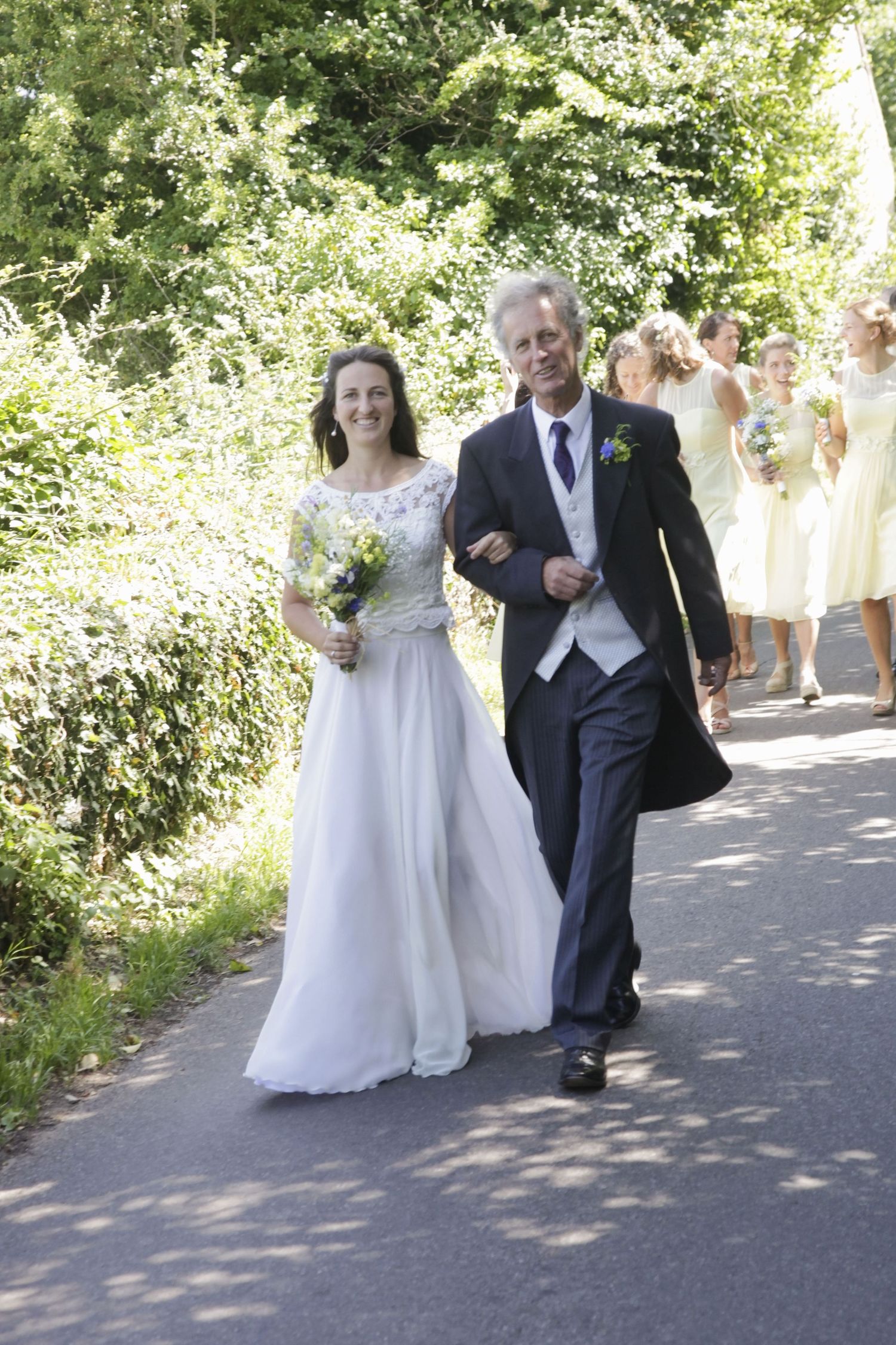 Wedding procession walking down a sunlit garden path with bridesmaids following behind.