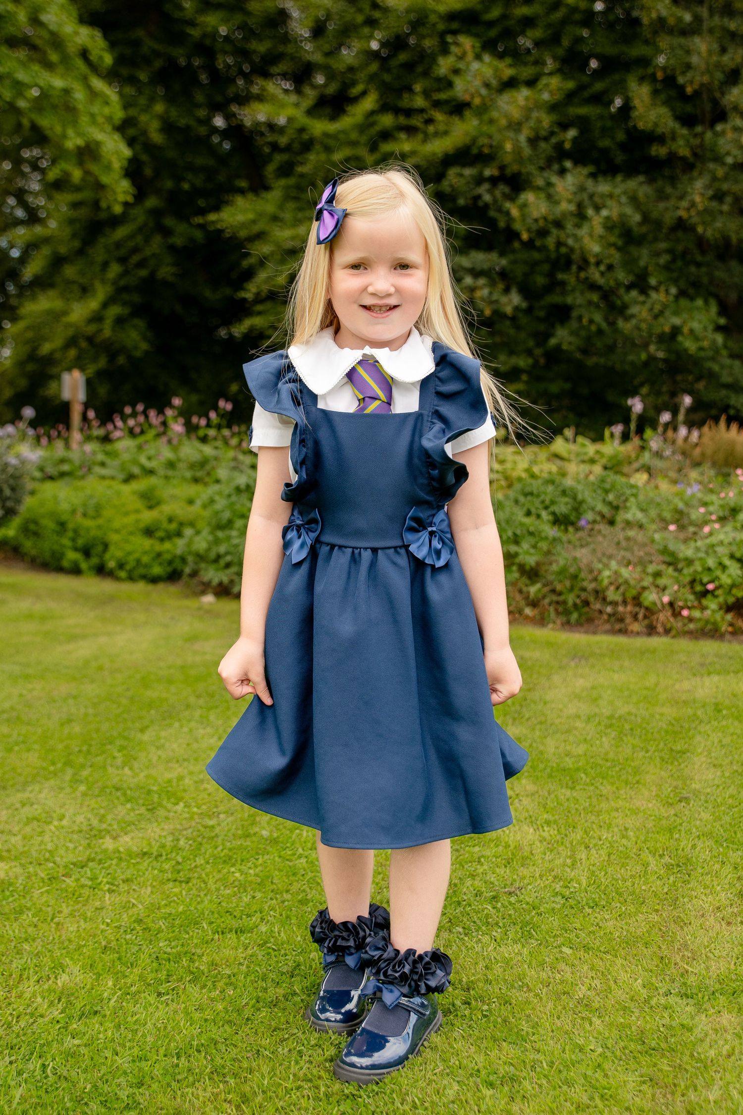 Young girl in navy school pinafore dress with ruffles, white shirt, and purple hair bow, smiling in a garden.