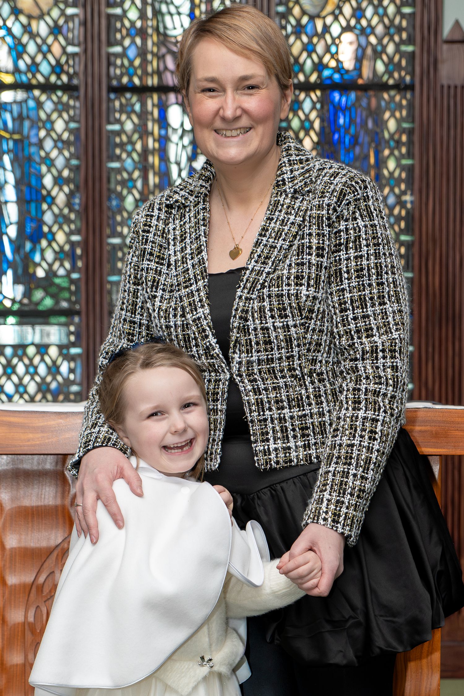 Woman in tweed jacket smiles with young girl in white dress inside a church with stained glass windows.