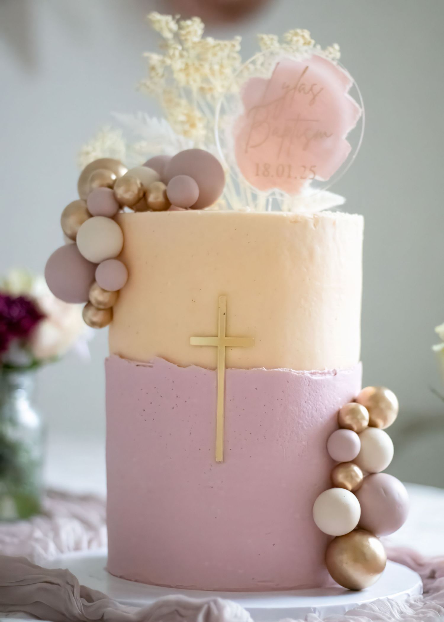 Two-tier pink and gold baptism cake with cross, balloon garland, and floral topper.