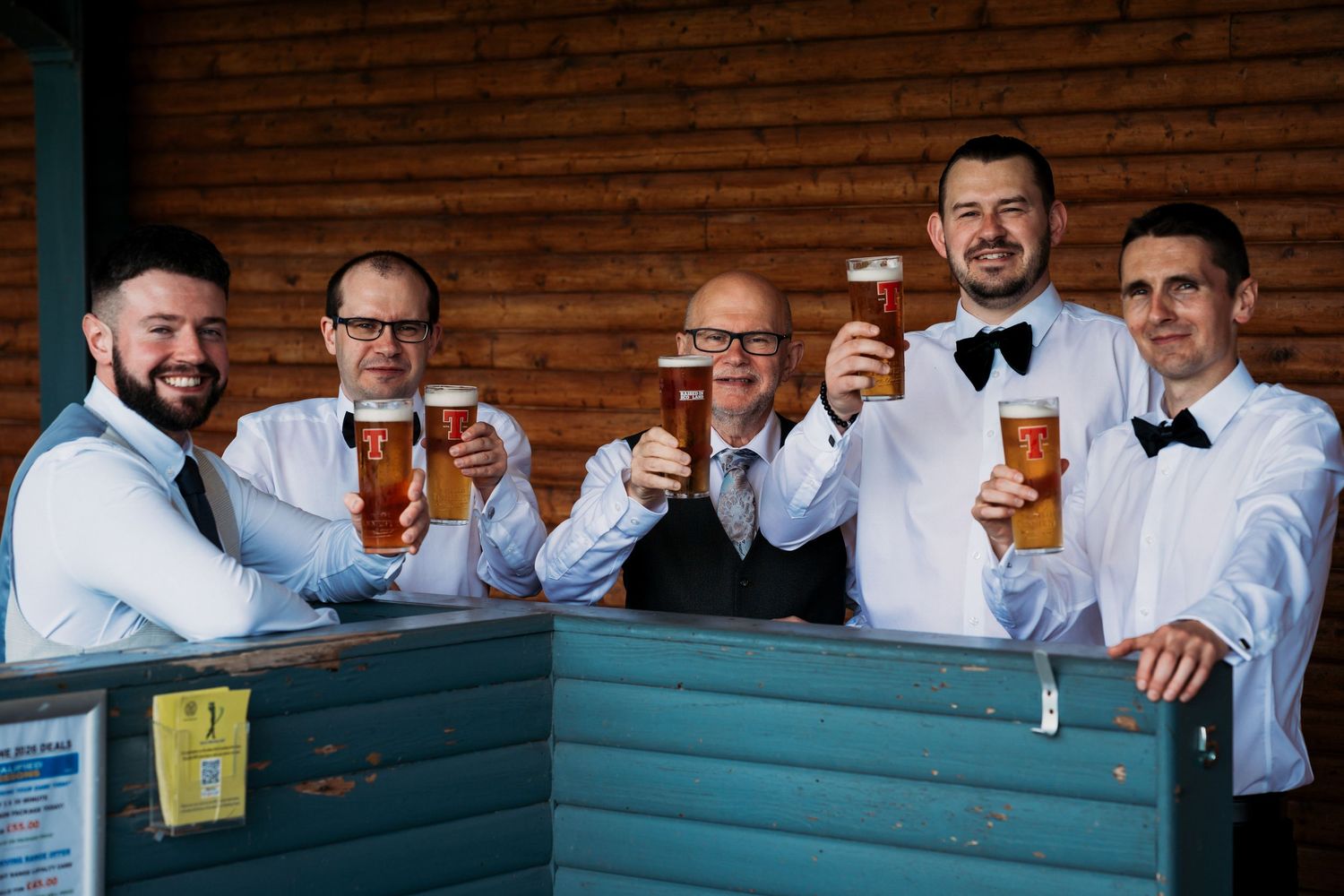 Five groomsmen in white shirts and bow ties toasting with beer glasses at a wedding celebration.