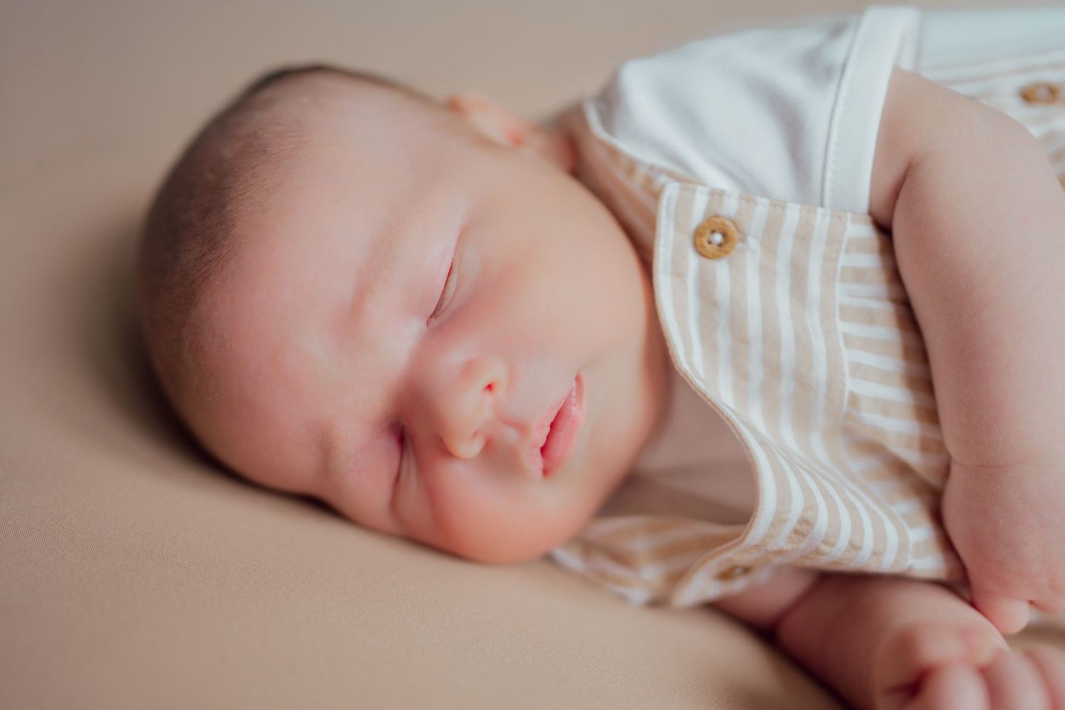 A peaceful newborn baby sleeping soundly, wearing a striped white and beige romper with suspenders.